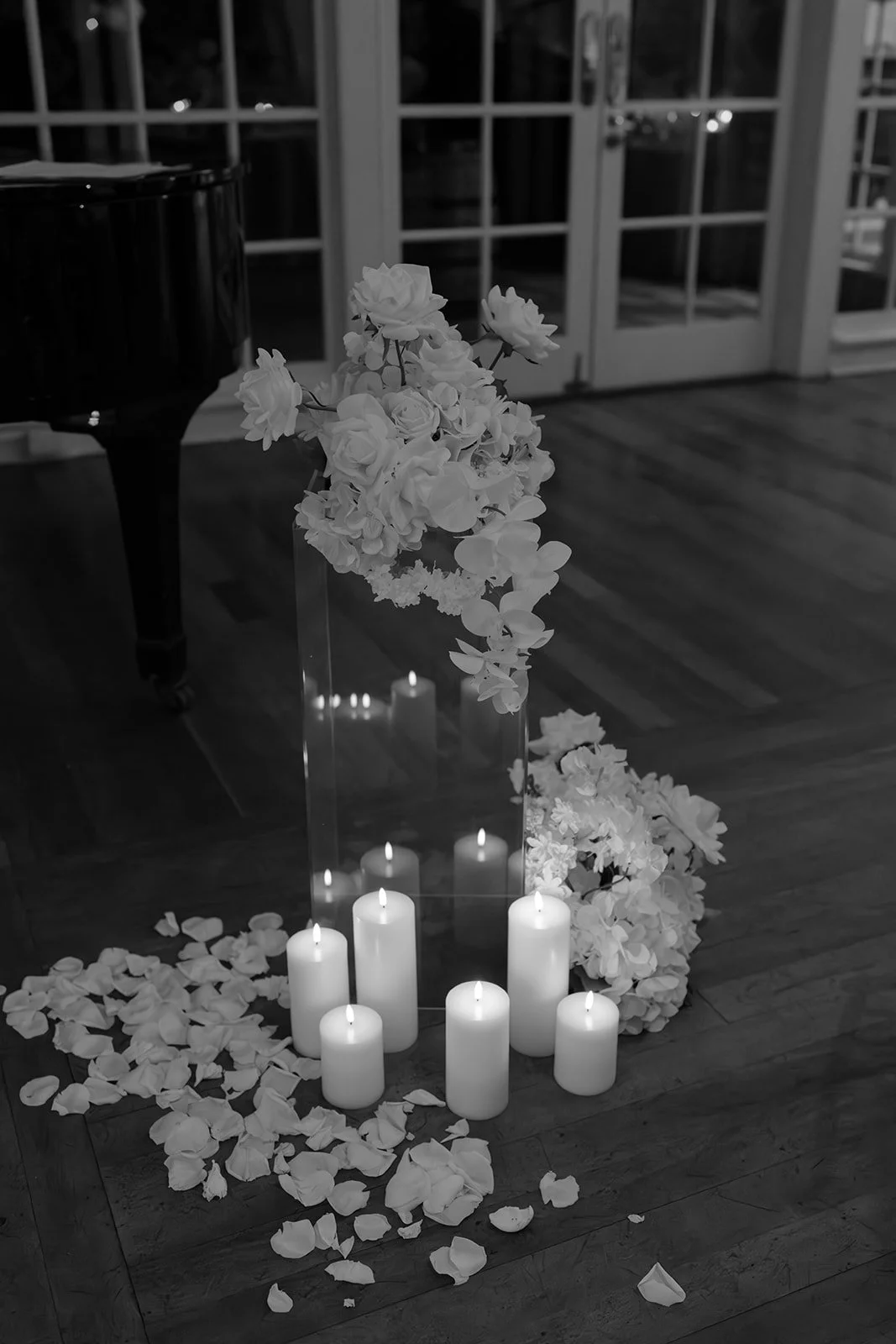 A floral arrangement with white flowers and white candles on a wooden floor, with a grand piano on the left and glass doors in the background.