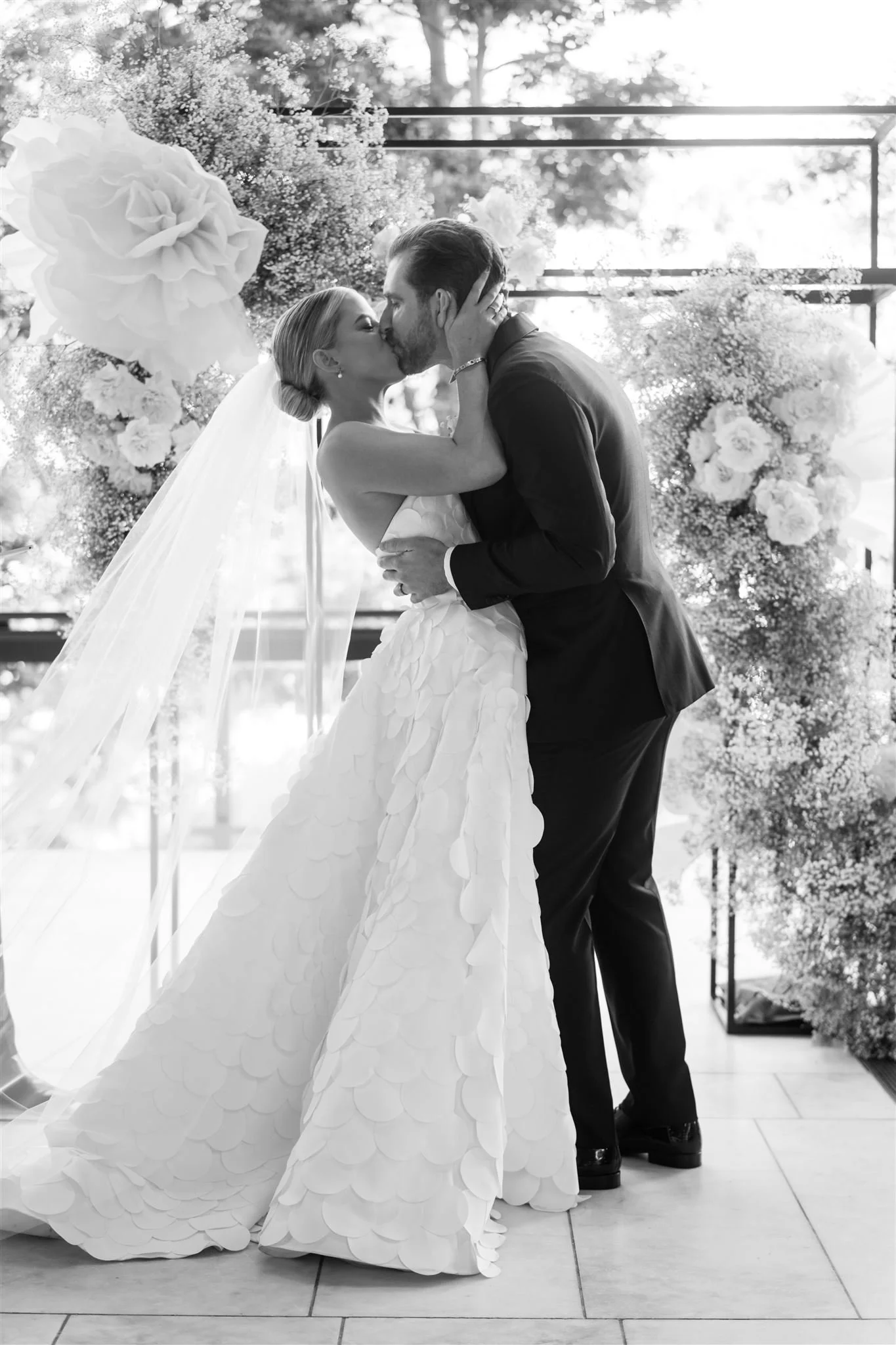 A bride and groom sharing a kiss during their wedding ceremony, standing in front of a floral arrangement with trees and a canopy in the background.