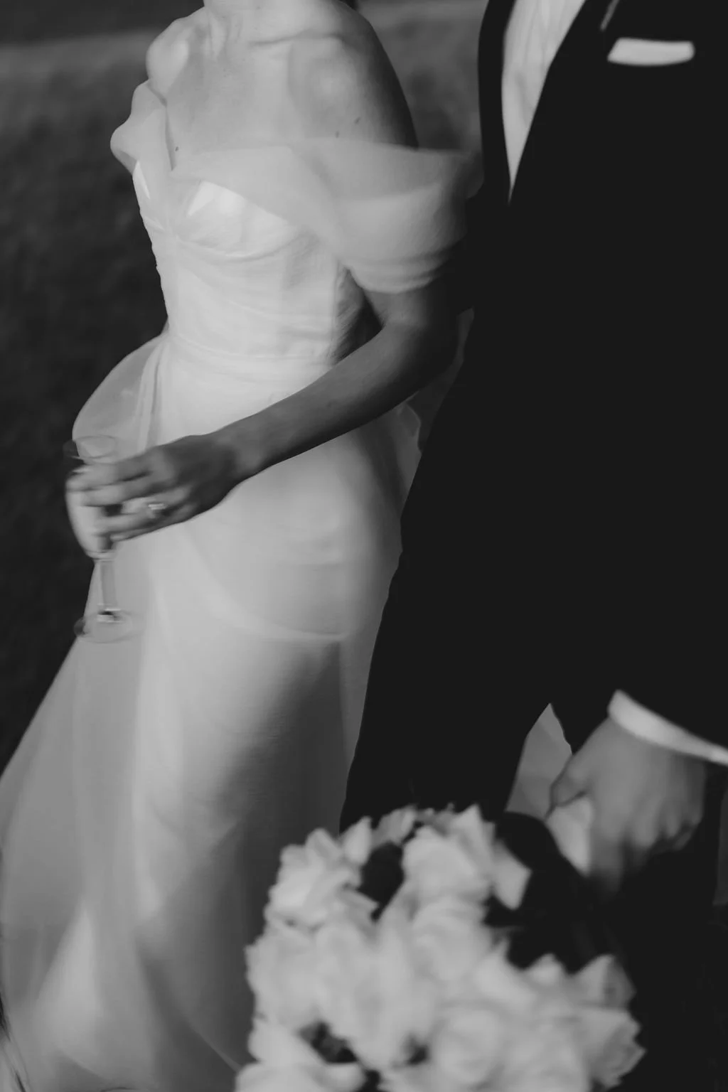 A bride and groom at a wedding reception, with the bride holding a glass and the groom resting his hand on a bouquet of flowers.