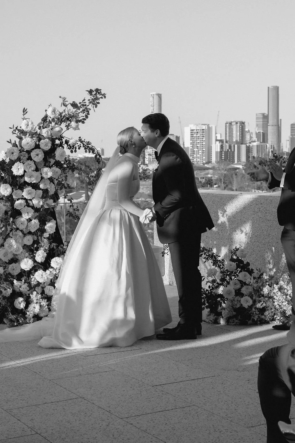 Black and white photo of a bride and groom kissing during their wedding ceremony on a rooftop with city skyline in the background, floral decoration on the side.