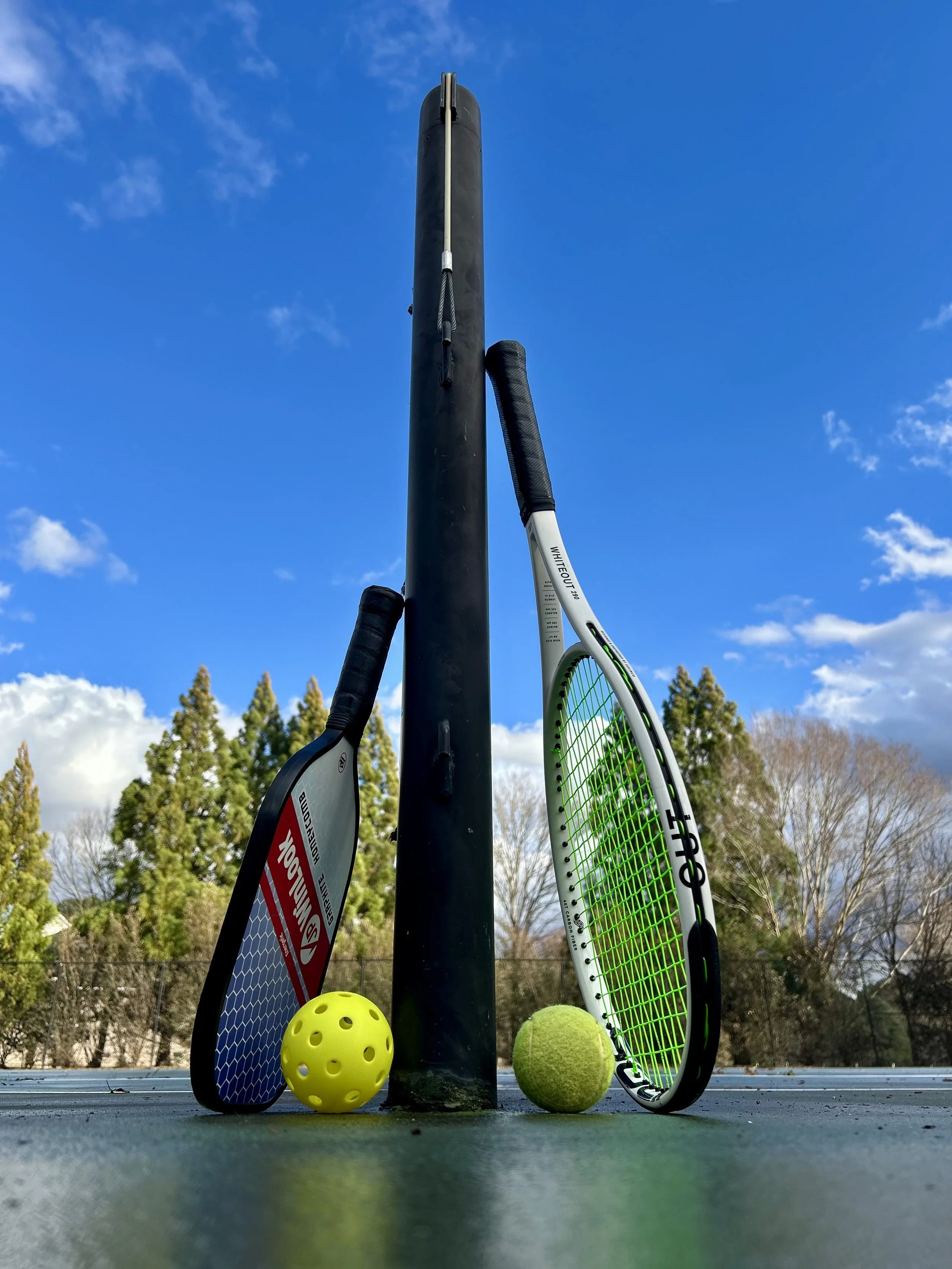 A pickleball paddle and ball lean against a net post on the left with a tennis racket and ball lean up on the right side of the net post with a blue sky and trees in the background