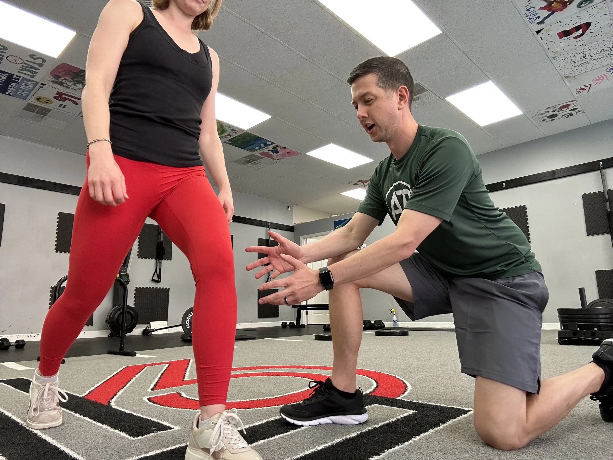 A physical therapist is assessing a patient's knee while they are lunging. The patient sustained a knee injury while playing pickleball.  The PT and patient are in a training gym with assorted dumbbells and weight plates in the background