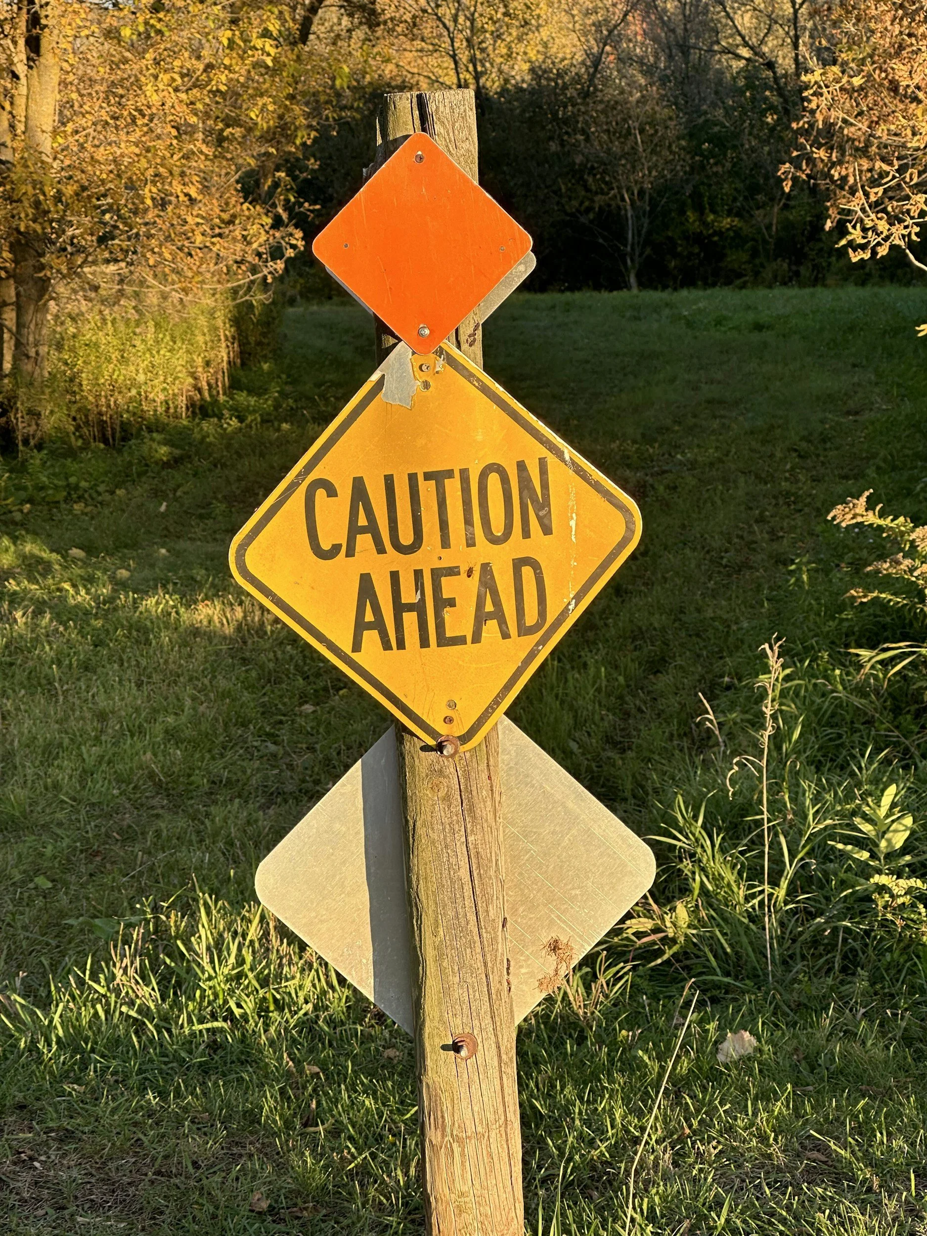 A yellow caution sign on a wooden post with an orange blank diamond-shaped sign on top, set outdoors in a grassy area with trees and bushes in the background.