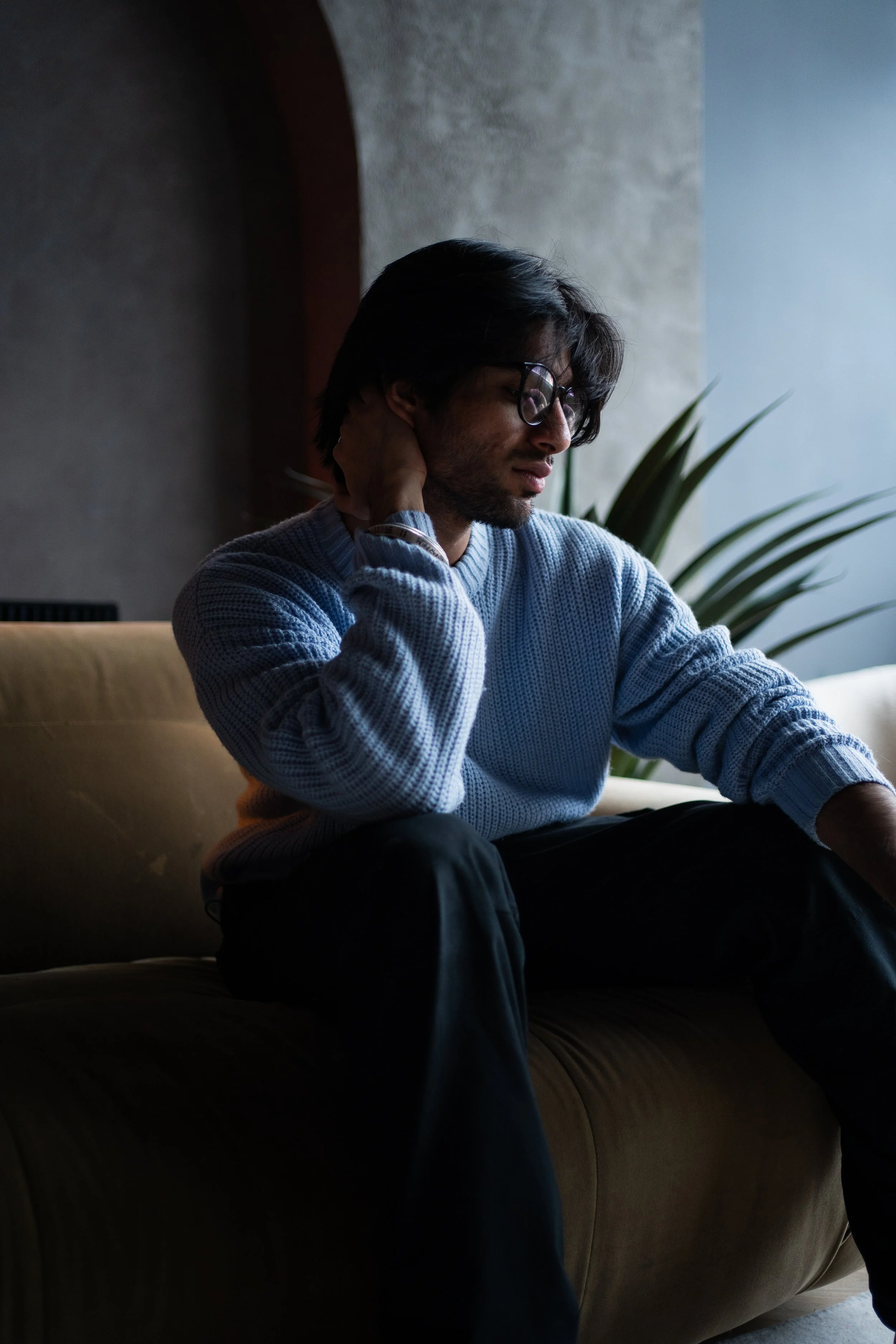 A young man with glasses, wearing a blue sweater, sitting on a sofa with a contemplative expression, resting his head on his hand, in a dimly lit room with a plant and a textured wall in the background.