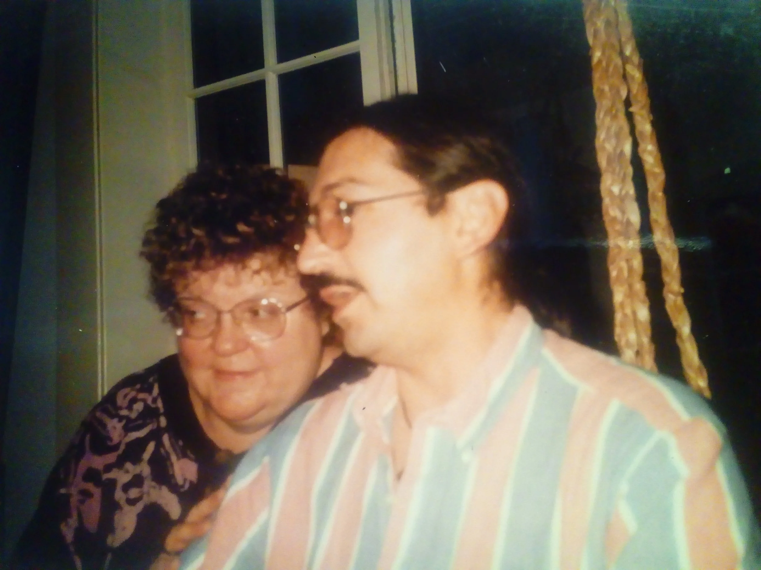 Two people sitting together indoors, one wearing glasses and a patterned top, the other with a mustache and striped shirt.