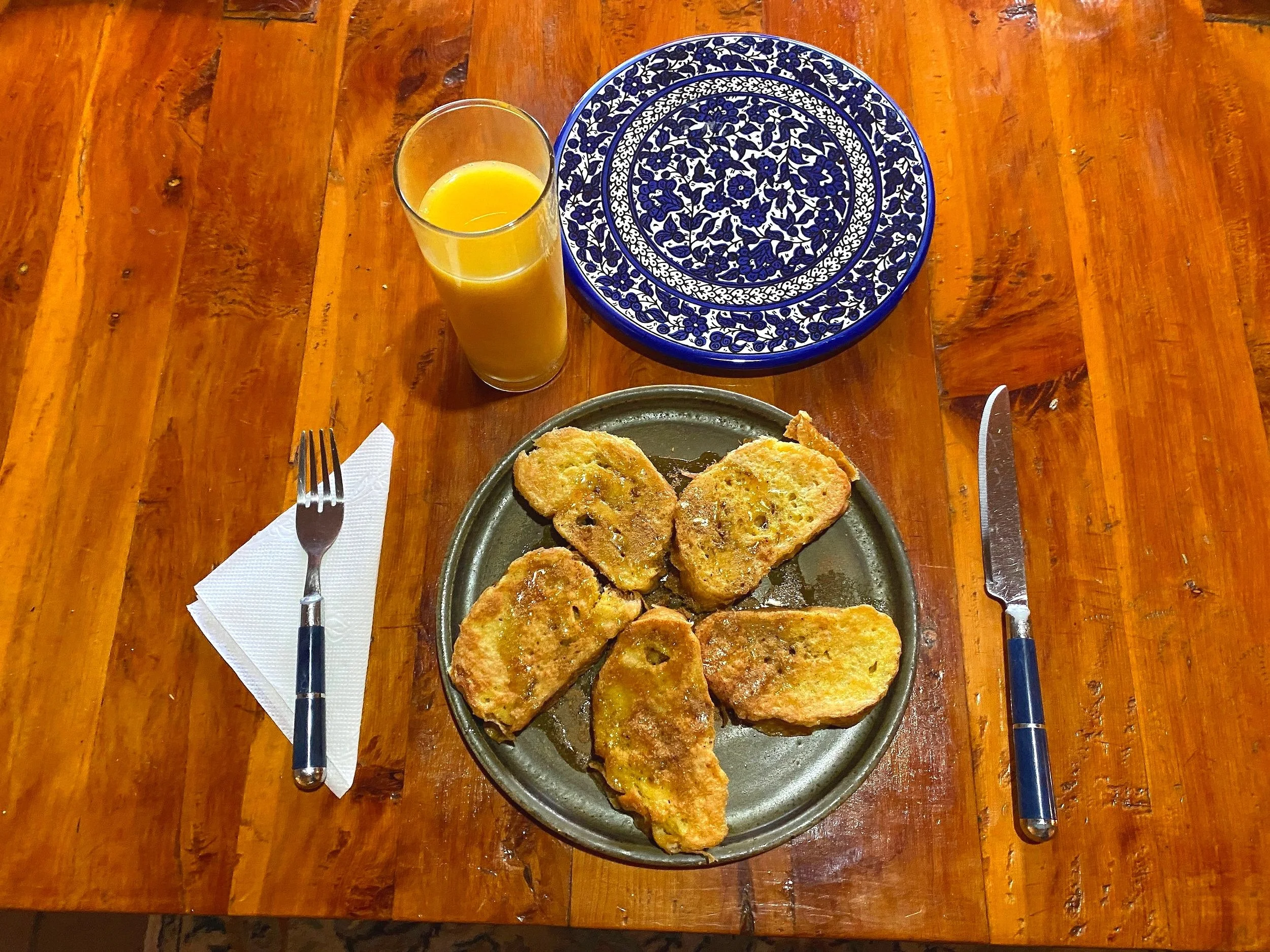 Plate of French toast with a glass of orange juice on a wooden table, accompanied by a fork, knife, and decorative plate.