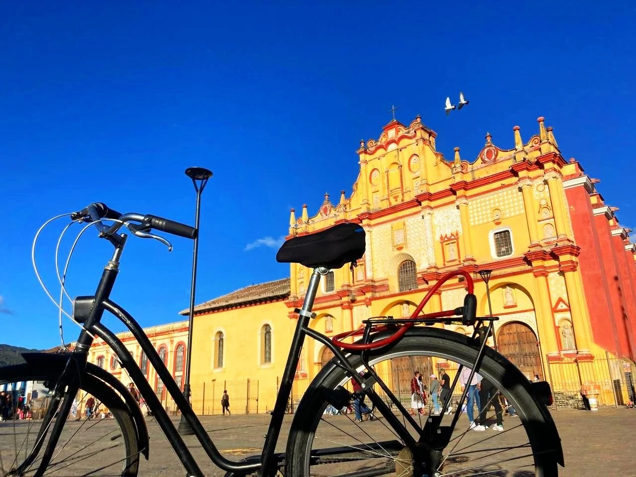 A bicycle parked in front of a vibrant yellow and red historical building with arched windows, under a clear blue sky. Two birds are flying above.