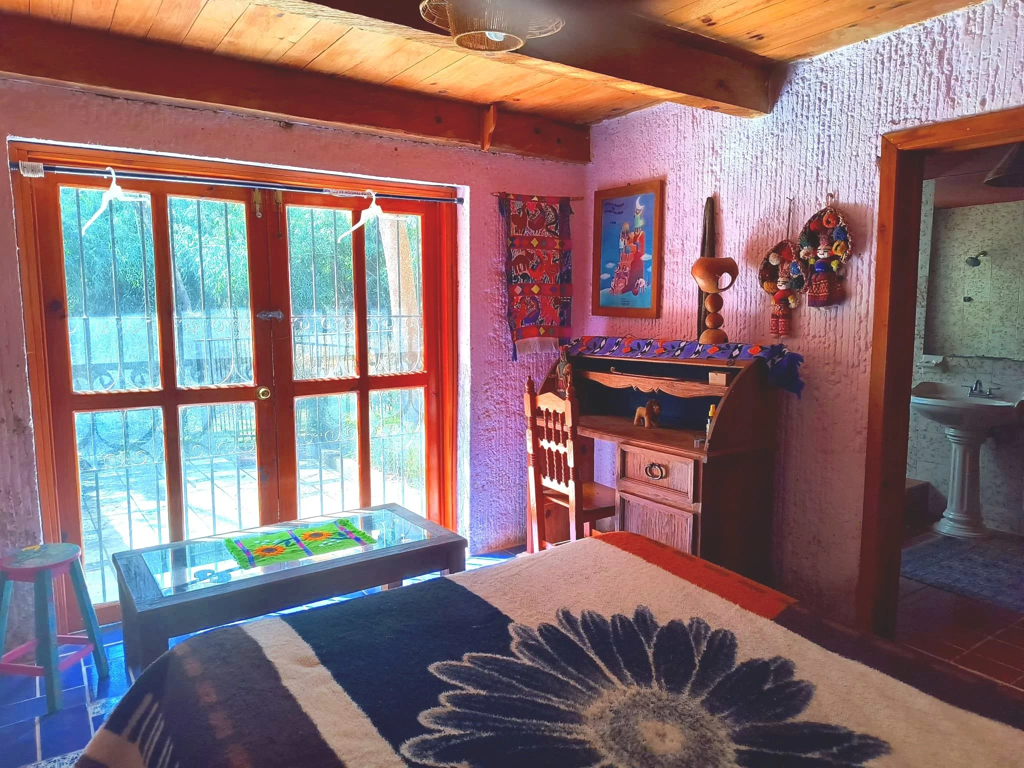 Cozy room with rustic decor, featuring a wooden desk, colorful wall art, and a bed with a floral blanket. There's a glass table, wooden stool, and a view of an outdoor area through large wooden doors.
