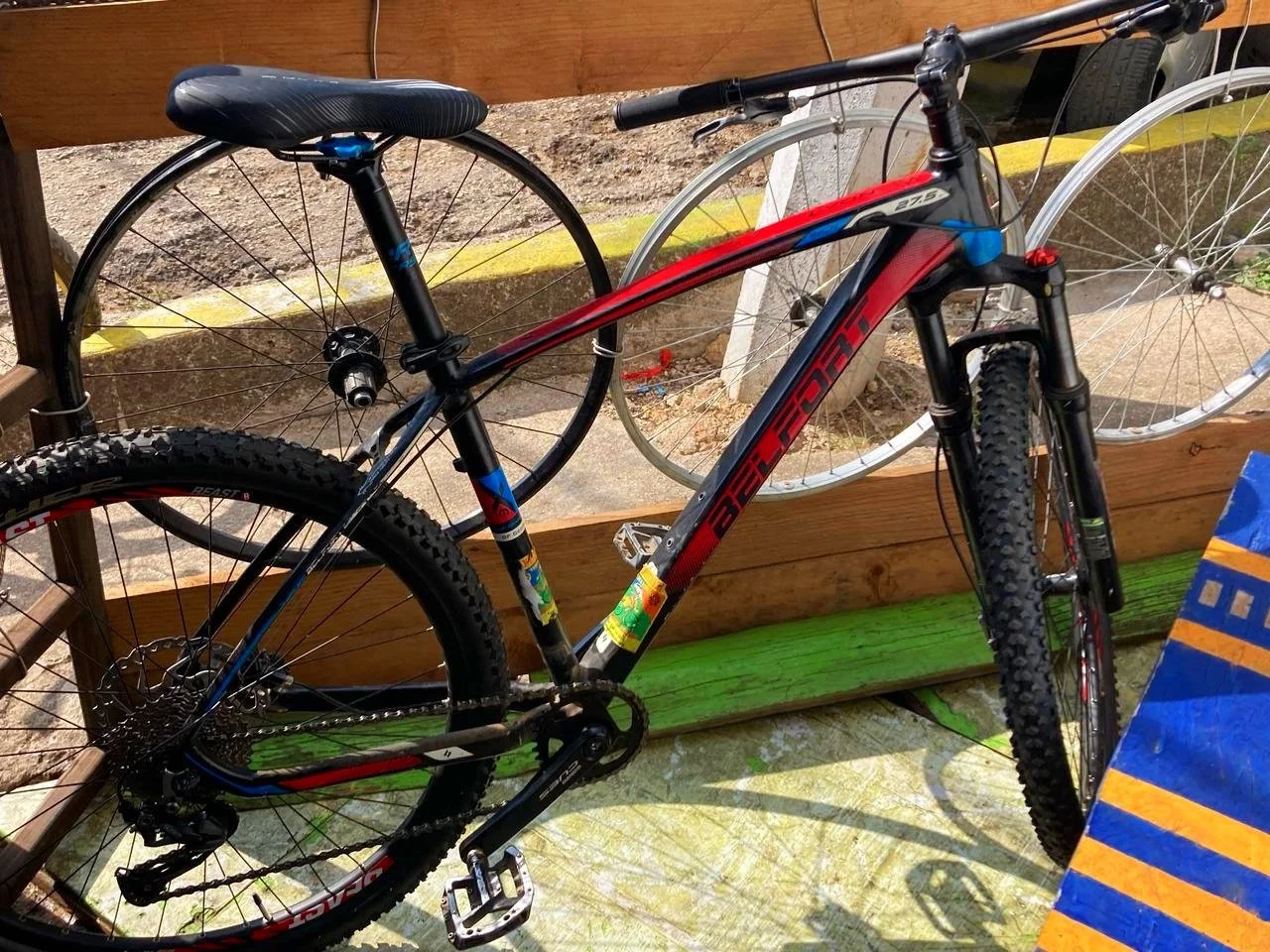 Mountain bike with a black and red frame, leaning against a wooden fence, with another wheel in the background.