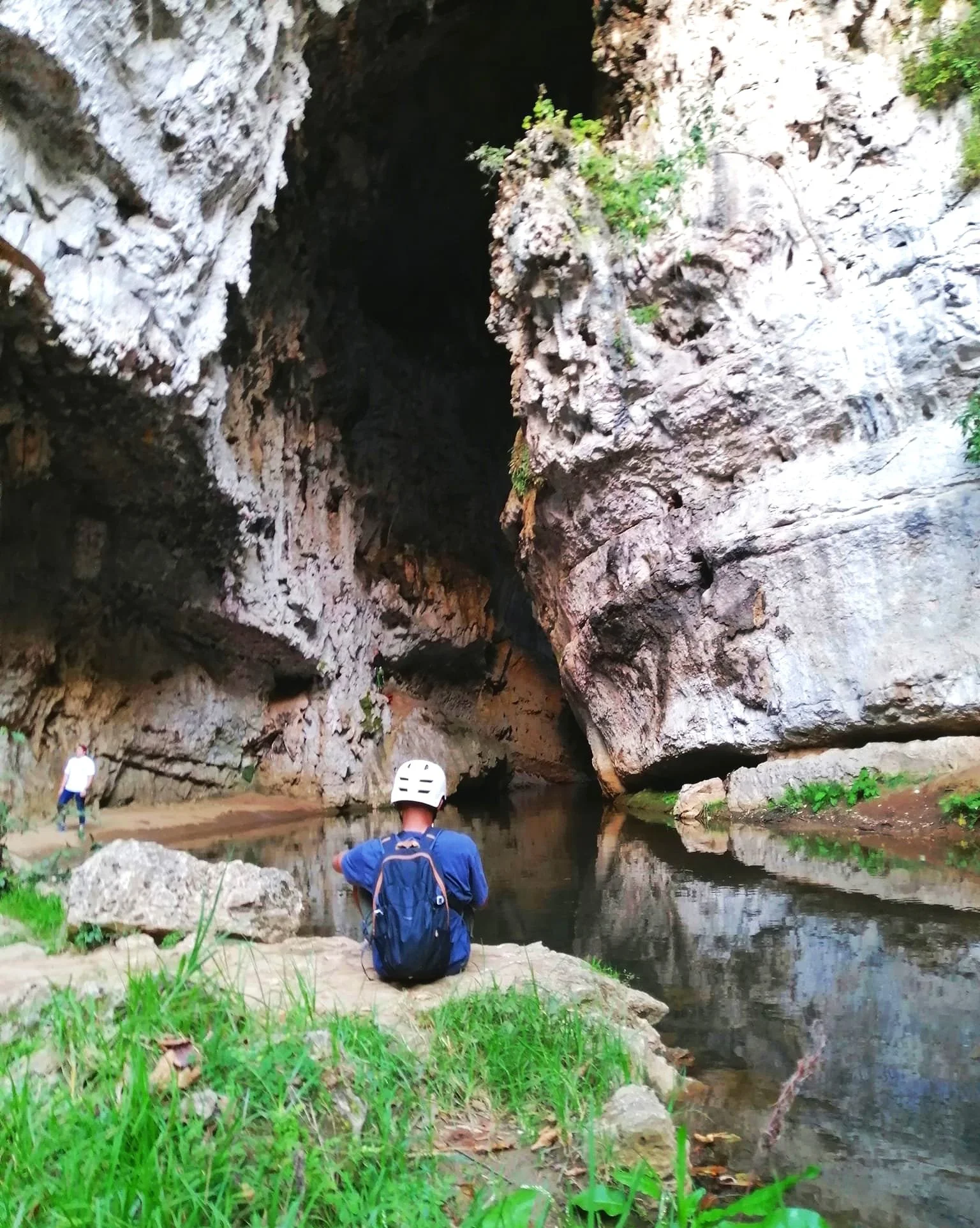 Person wearing helmet and backpack sitting near a cave entrance with rocky walls and water, surrounded by greenery.