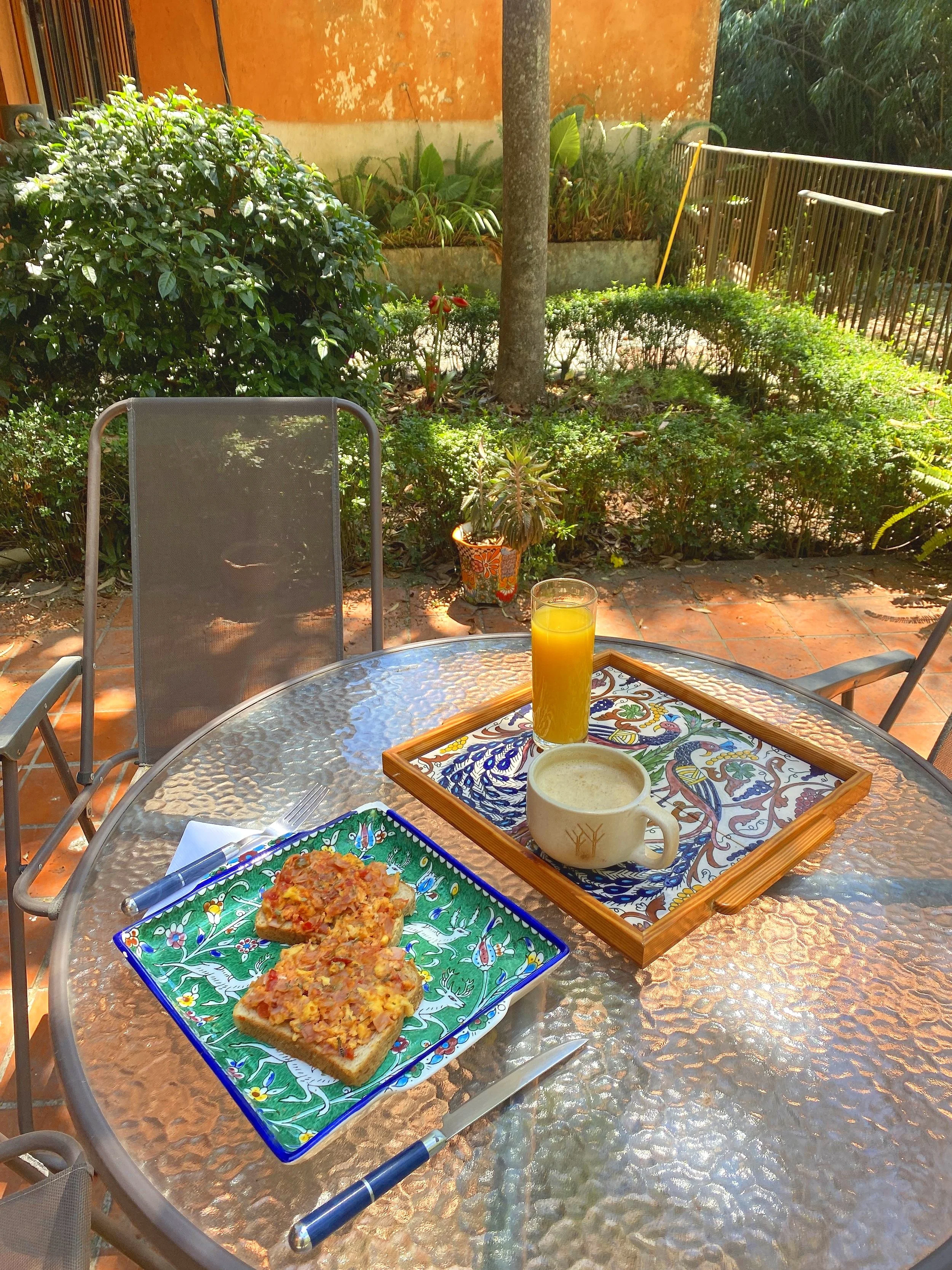 Outdoor table with a glass plate of toast topped with onions and cheese, a glass of orange juice, a cappuccino on a decorative tray, surrounded by garden greenery.
