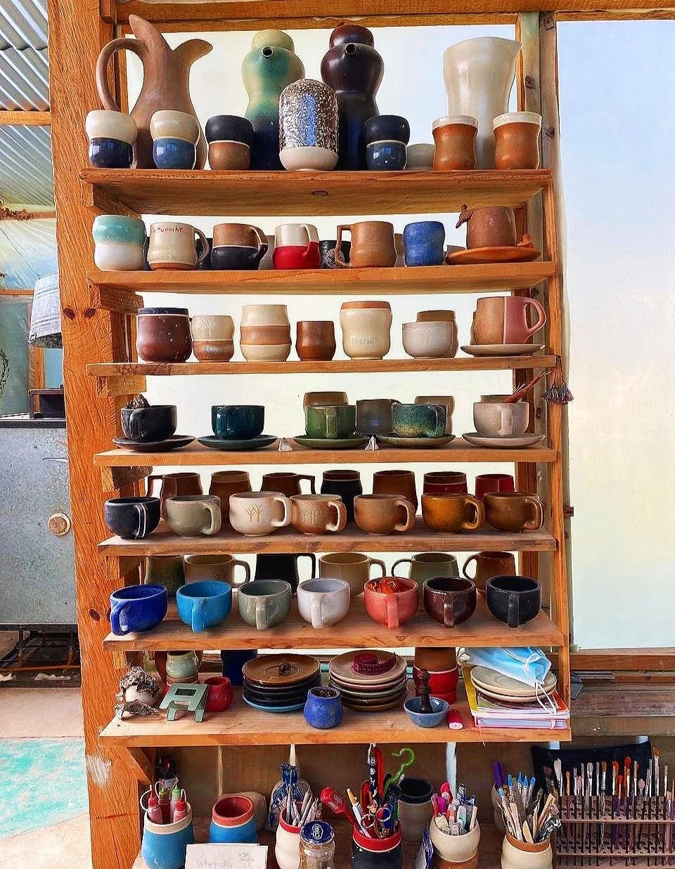 Wooden shelves displaying various colorful ceramic mugs, cups, pots, and art supplies in a studio setting.