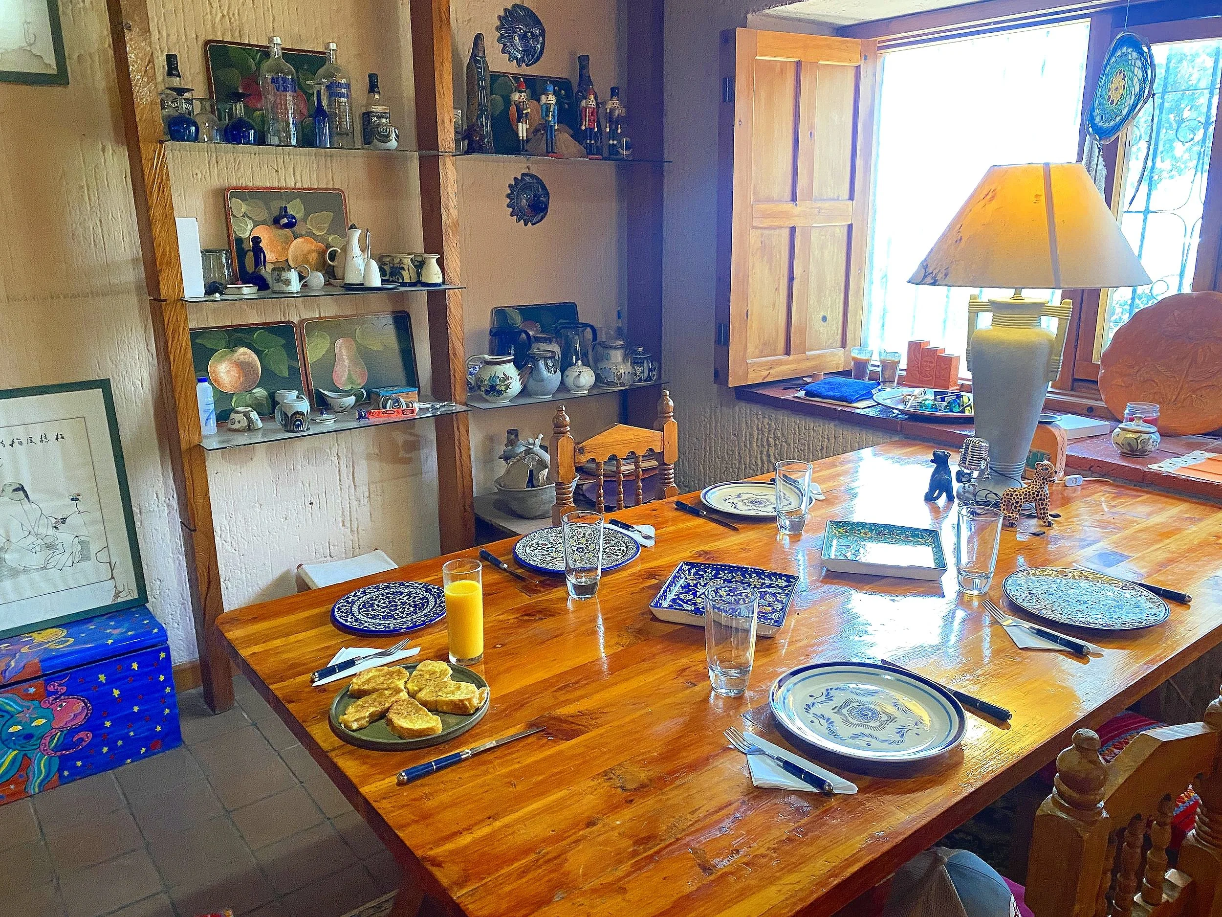 Dining room with wooden table set for a meal, featuring decorative plates, glassware, and a plate of orange slices. Shelving unit with various knick-knacks, bottles, and ceramics. Natural light from a window on the side.