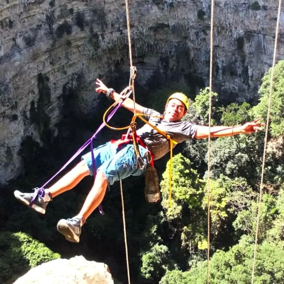 Person wearing a helmet and harness, rappelling in a canyon with trees and rocky cliff in the background.
