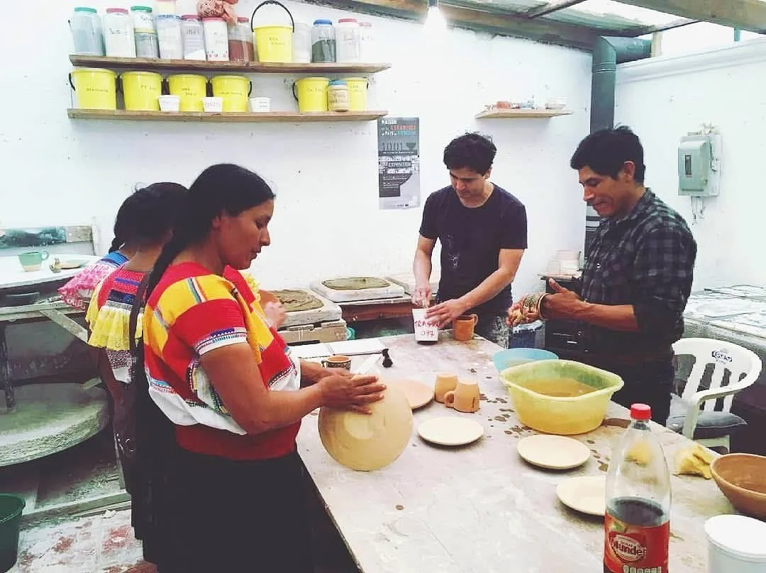 Group of people making pottery in a workshop, with shelves of containers in the background. Participants are shaping and handling clay objects on a table.
