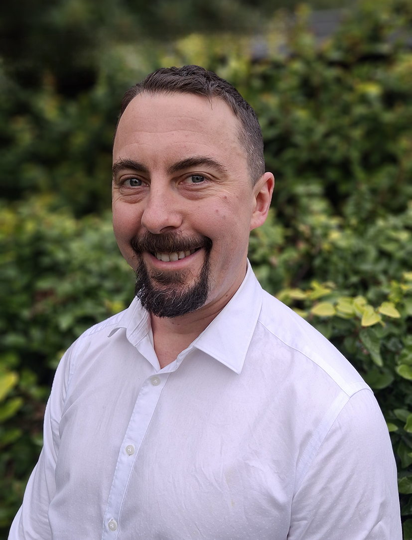 Portrait of a smiling man with short dark hair and a trimmed beard, wearing a white collared shirt outdoors against a leafy green background.