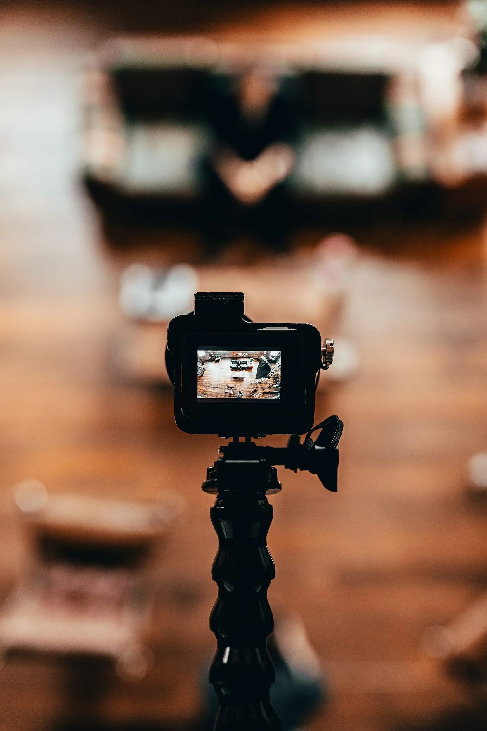 A camera on a tripod capturing a bird's-eye view of an indoor setting with wooden flooring and various furniture pieces, including tables and chairs, with a person sitting in the background.