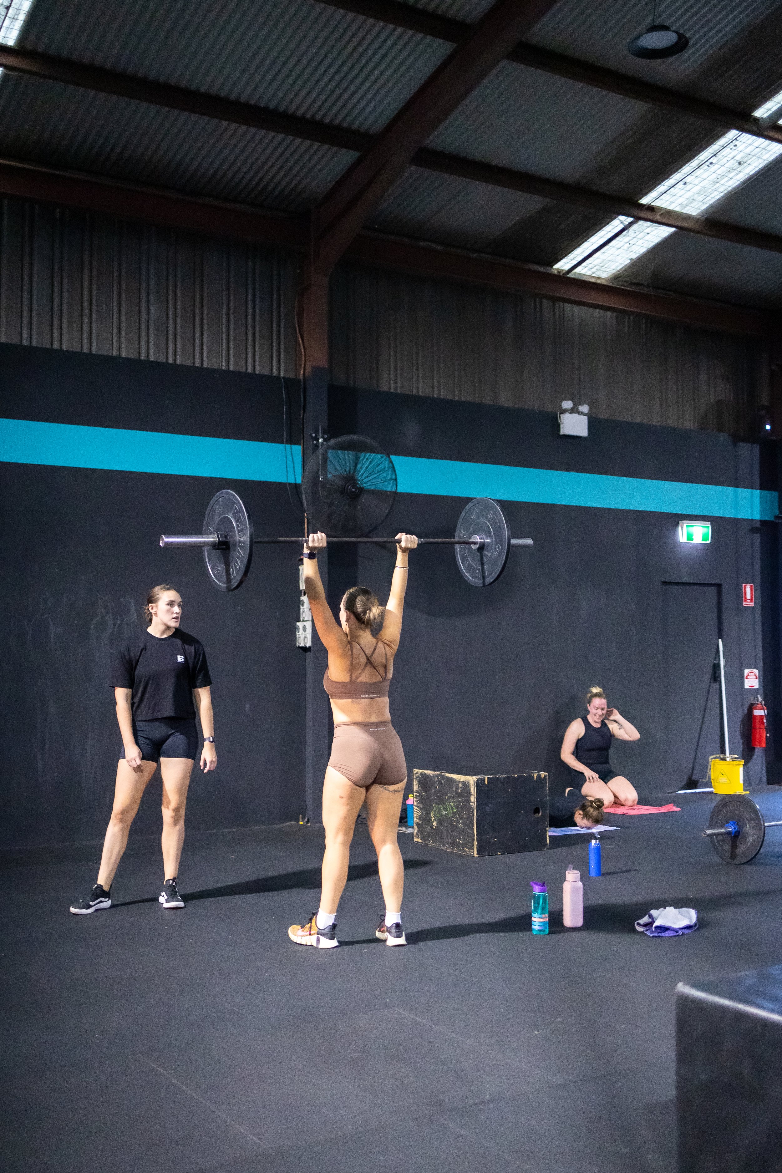 Woman lifting a barbell overhead in a gym with two women sitting on mats in the background.