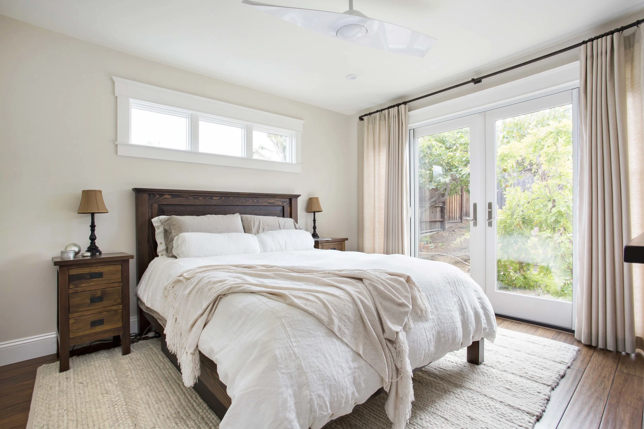 A bedroom with a wooden bed frame, white bedding, beige blanket, two matching nightstands with lamps, beige curtains, a sliding glass door, and a window overhead, overlooking a backyard with trees.
