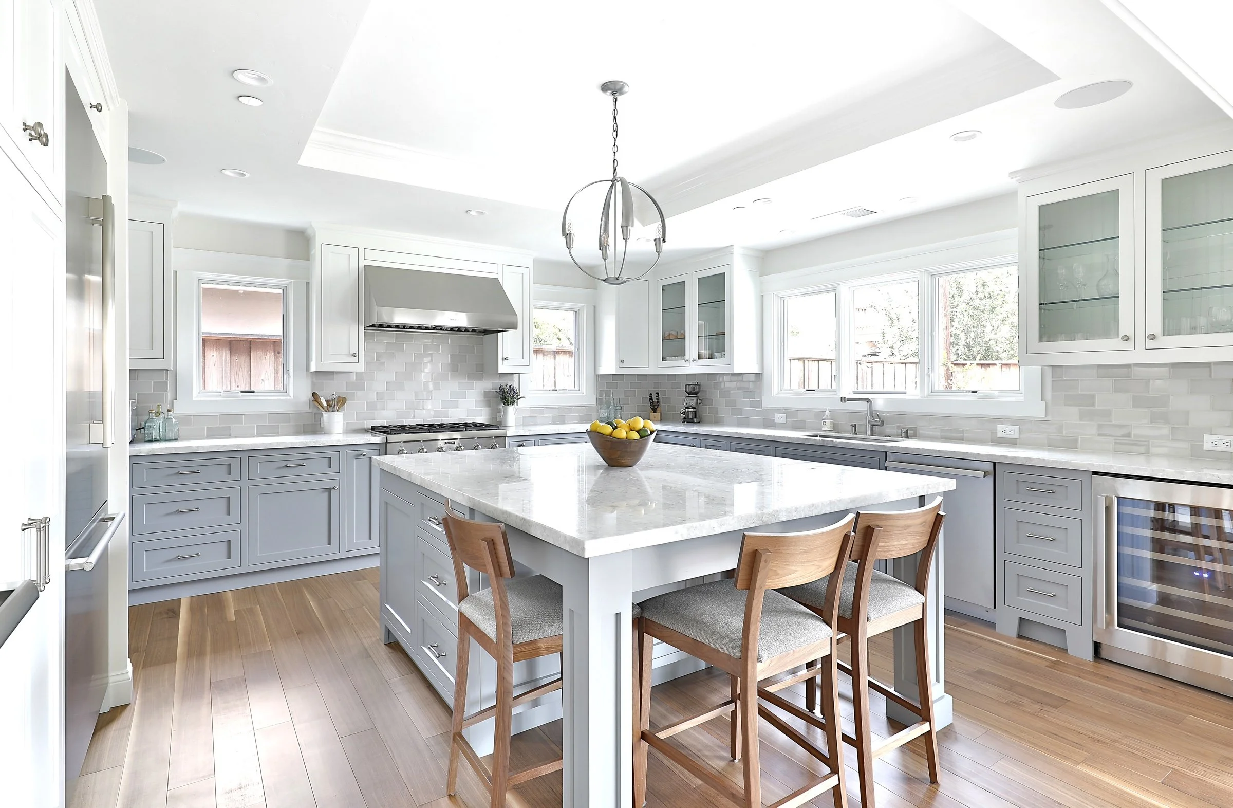 Bright modern kitchen with white cabinets, grey lower cabinets, a central island with a white marble countertop, three wooden chairs, and multiple large windows with natural light.