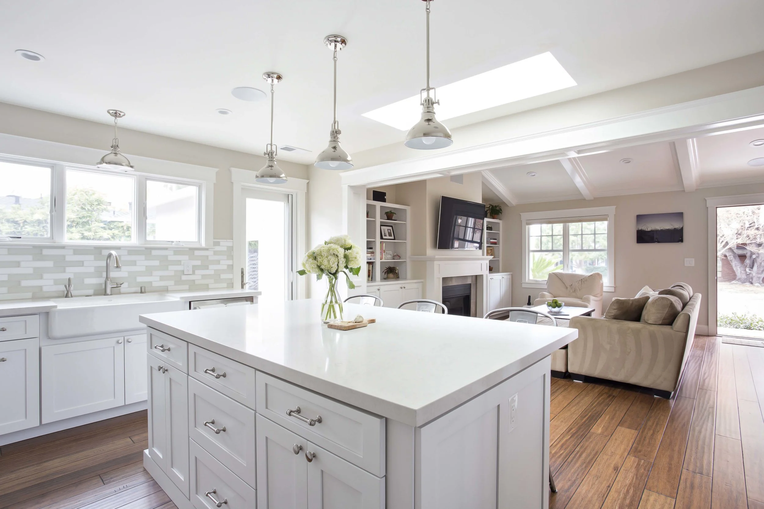 A bright and spacious open-concept kitchen and living room with white cabinets, a large island with a vase of white flowers, a fireplace, a wall-mounted TV, beige sofas, and large windows letting in natural light.