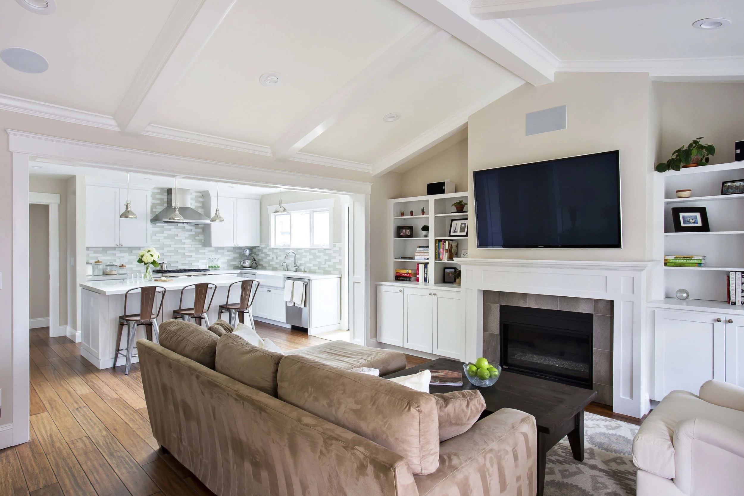 Living room with beige couch, white fireplace, mounted flat-screen TV, built-in white bookshelves, and a dark wood coffee table with a bowl of green apples. Open kitchen with white cabinets, gray backsplash, and breakfast bar with three chairs.