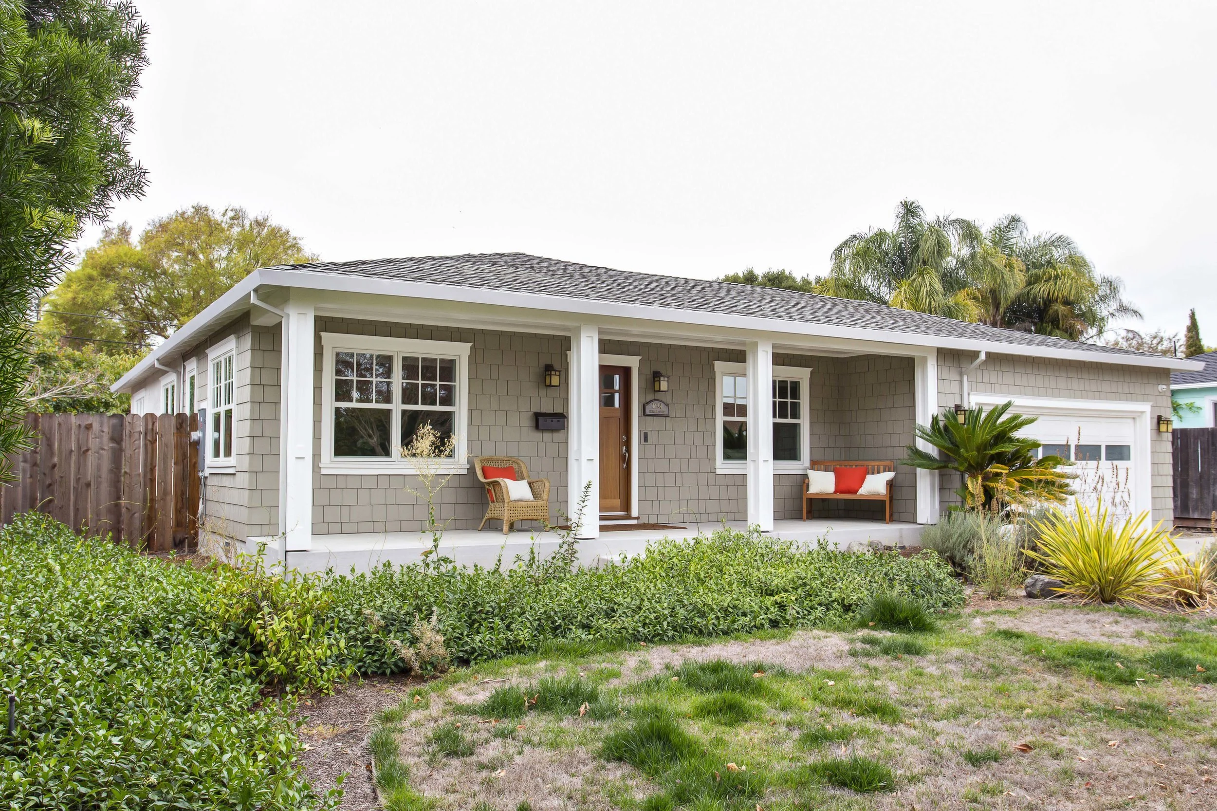 Front view of a single-story house with a covered porch, beige siding, white trim, and a brown front door. There are two outdoor chairs and a bench on the porch. The yard has green shrubs, grass patches, and desert plants. Tall trees and a cloudy sky