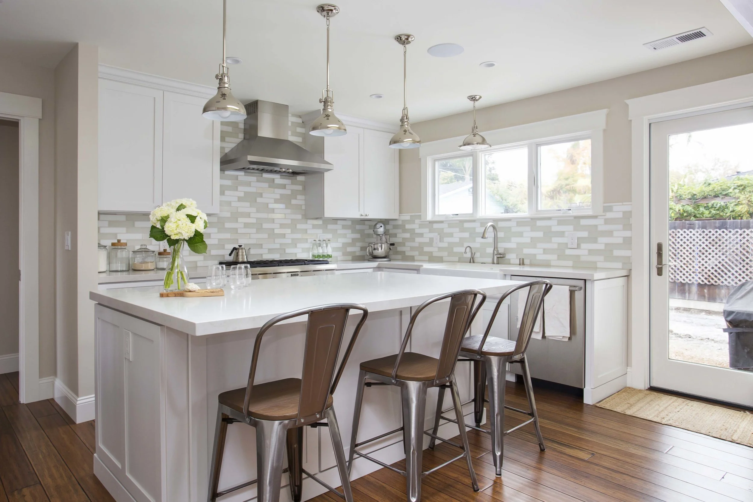 Modern white kitchen with a large island, four stools, pendant lights, a window over the sink, and a glass door leading outside.