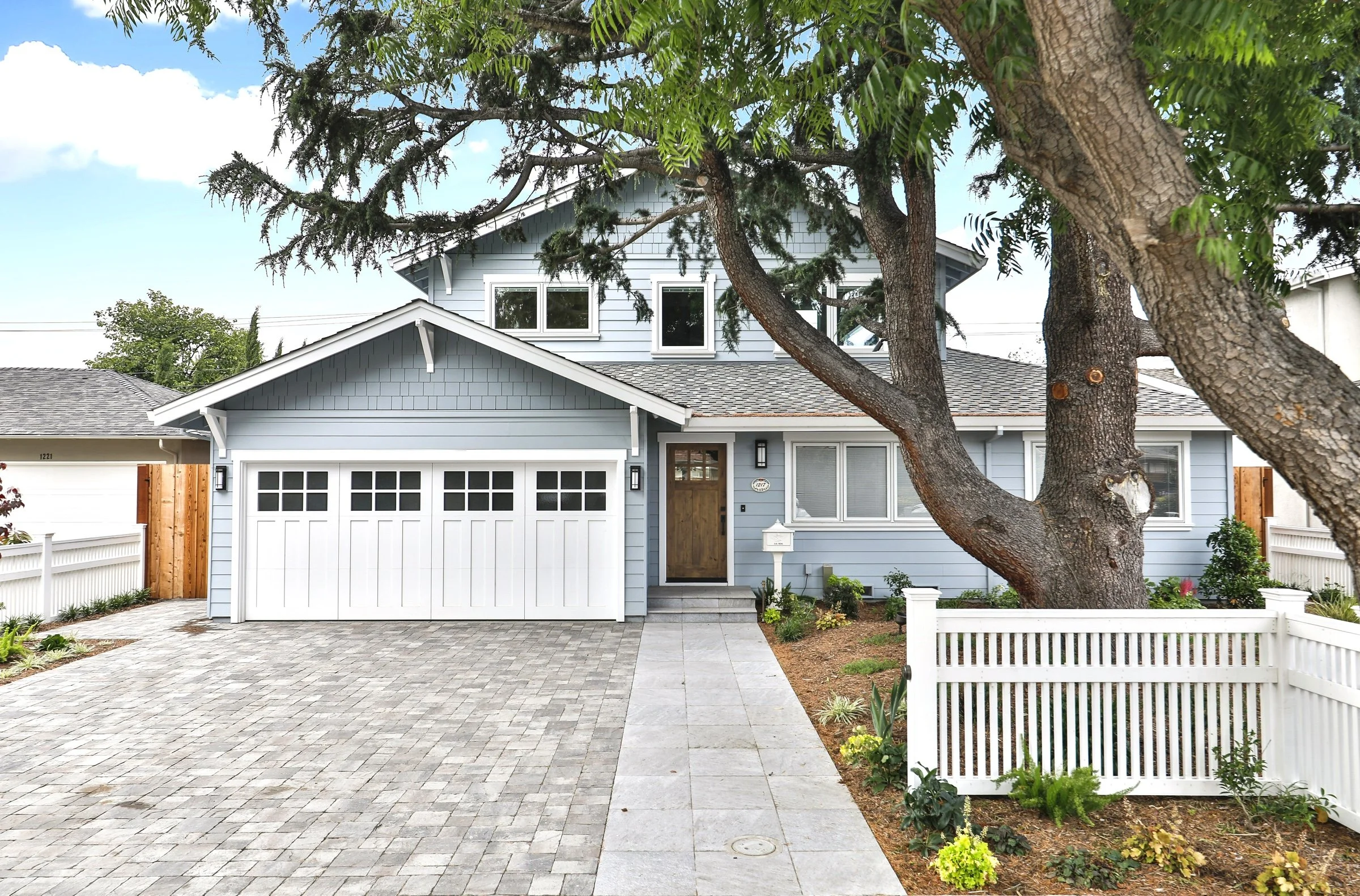 A two-story blue house with white trim, a wooden front door, and a white garage door. There is a large tree in front, with branches extending over the house. The front yard has a paved driveway, a concrete walkway, and a small garden with green plant