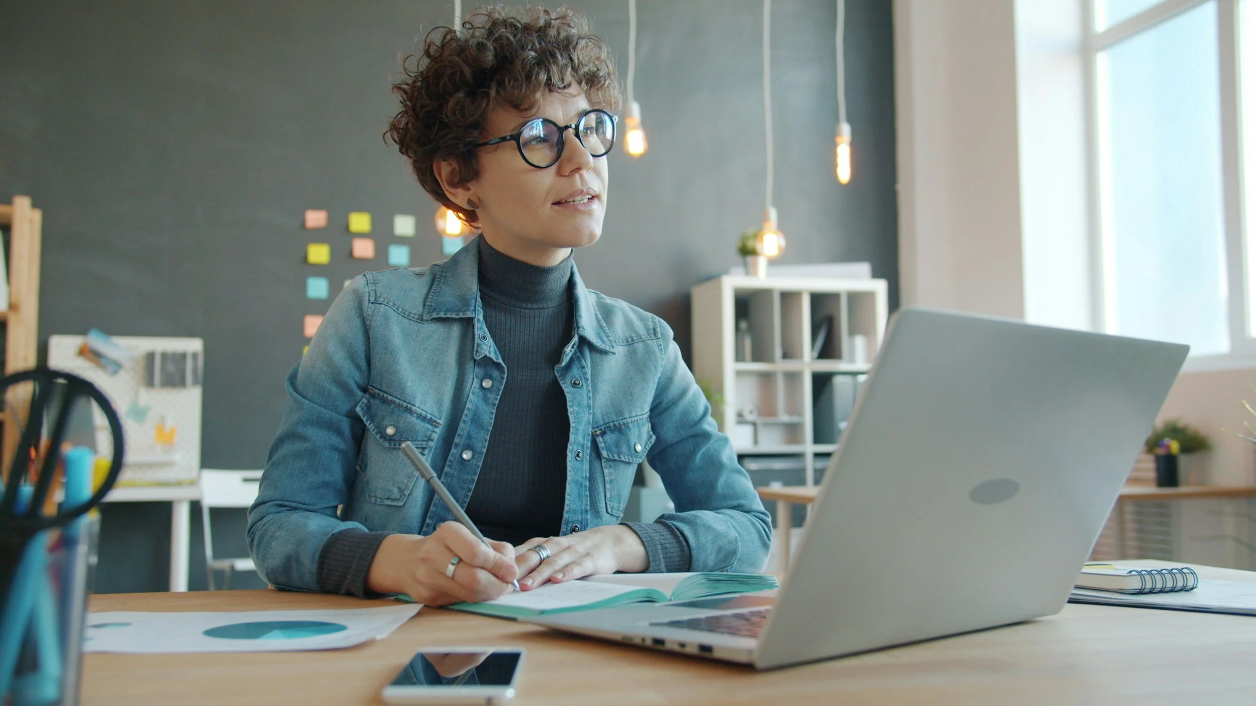 A focused woman with glasses taking notes while reviewing financial data on a laptop, representing monthly bookkeeping management.