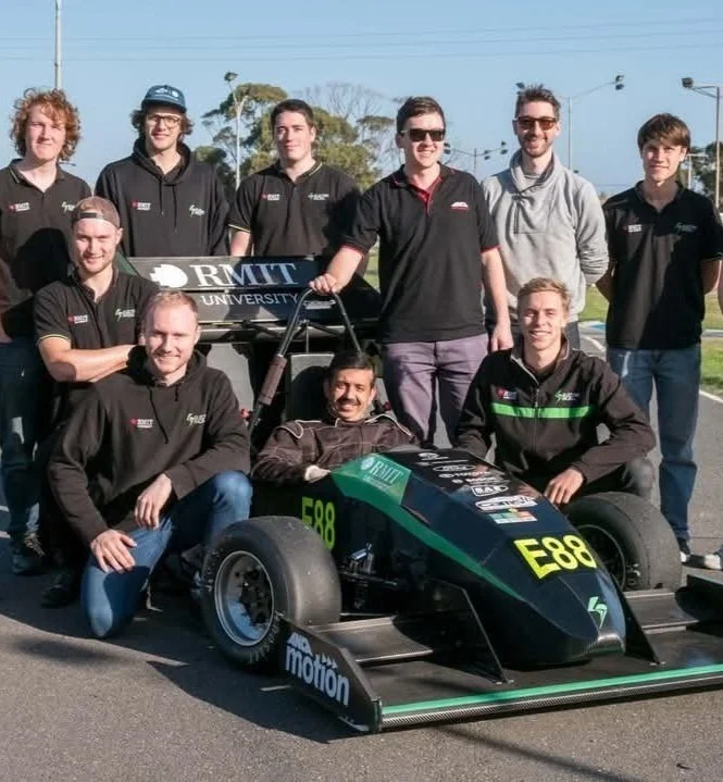 A group of people standing and crouching around a small racing car labeled E88 and RMIT University, outdoors on a track.