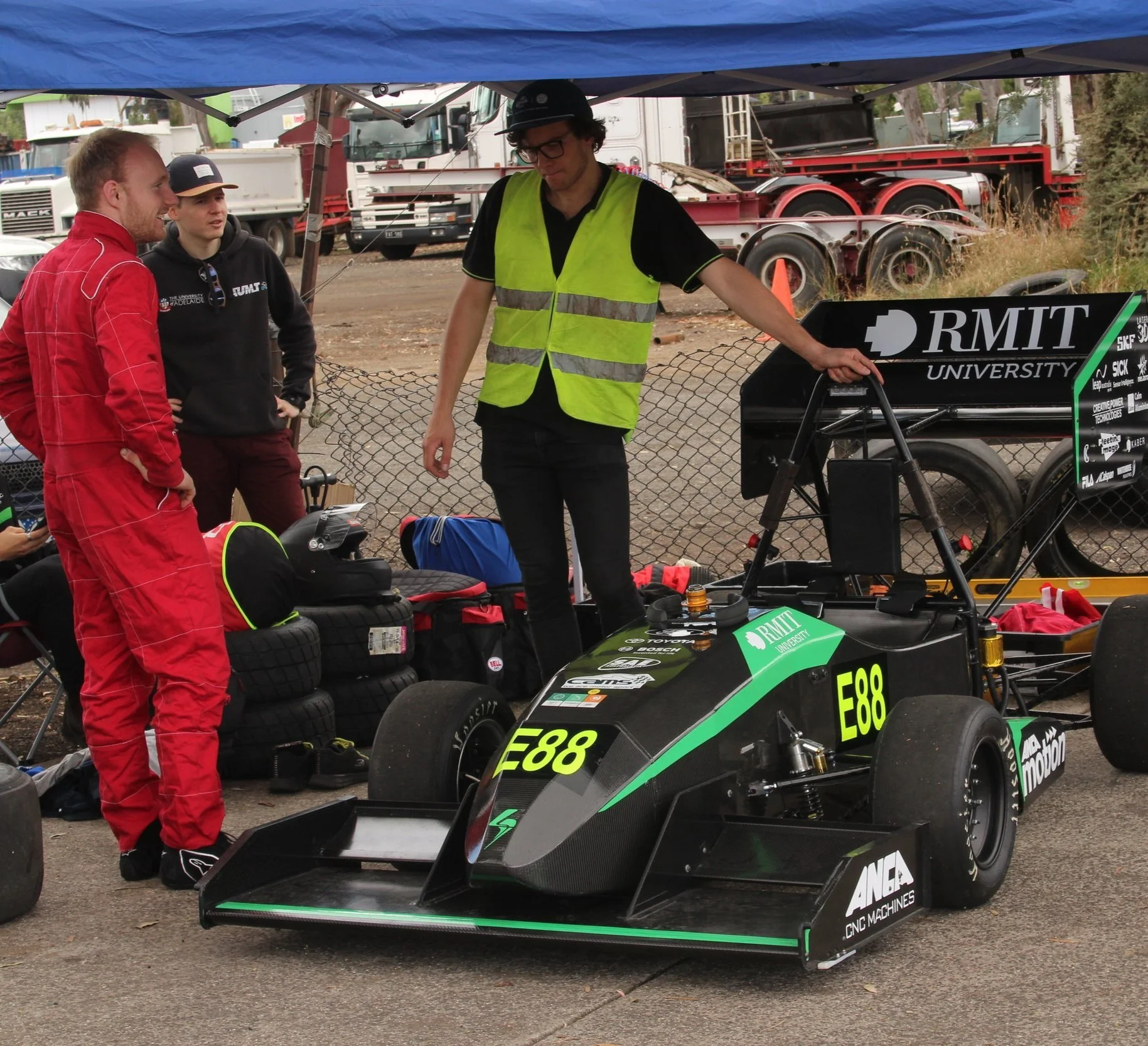 Three people discussing next to a race car in a pit area, with one wearing a red racing suit, another in a reflective vest, and a third in casual attire.