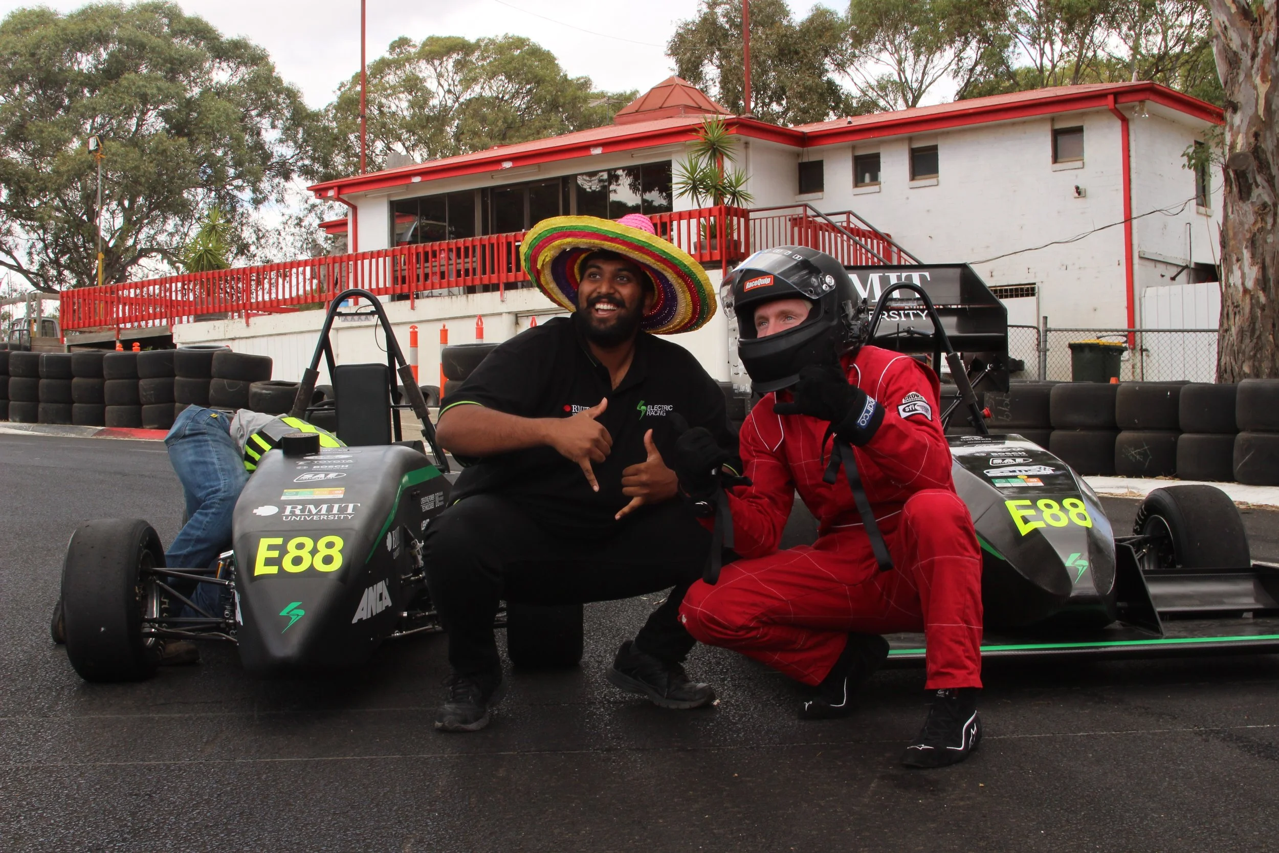 Two people smiling at a race track; one wearing a racing suit and helmet, the other wearing a sombrero and black shirt, next to a Formula Student car with 'E88'"