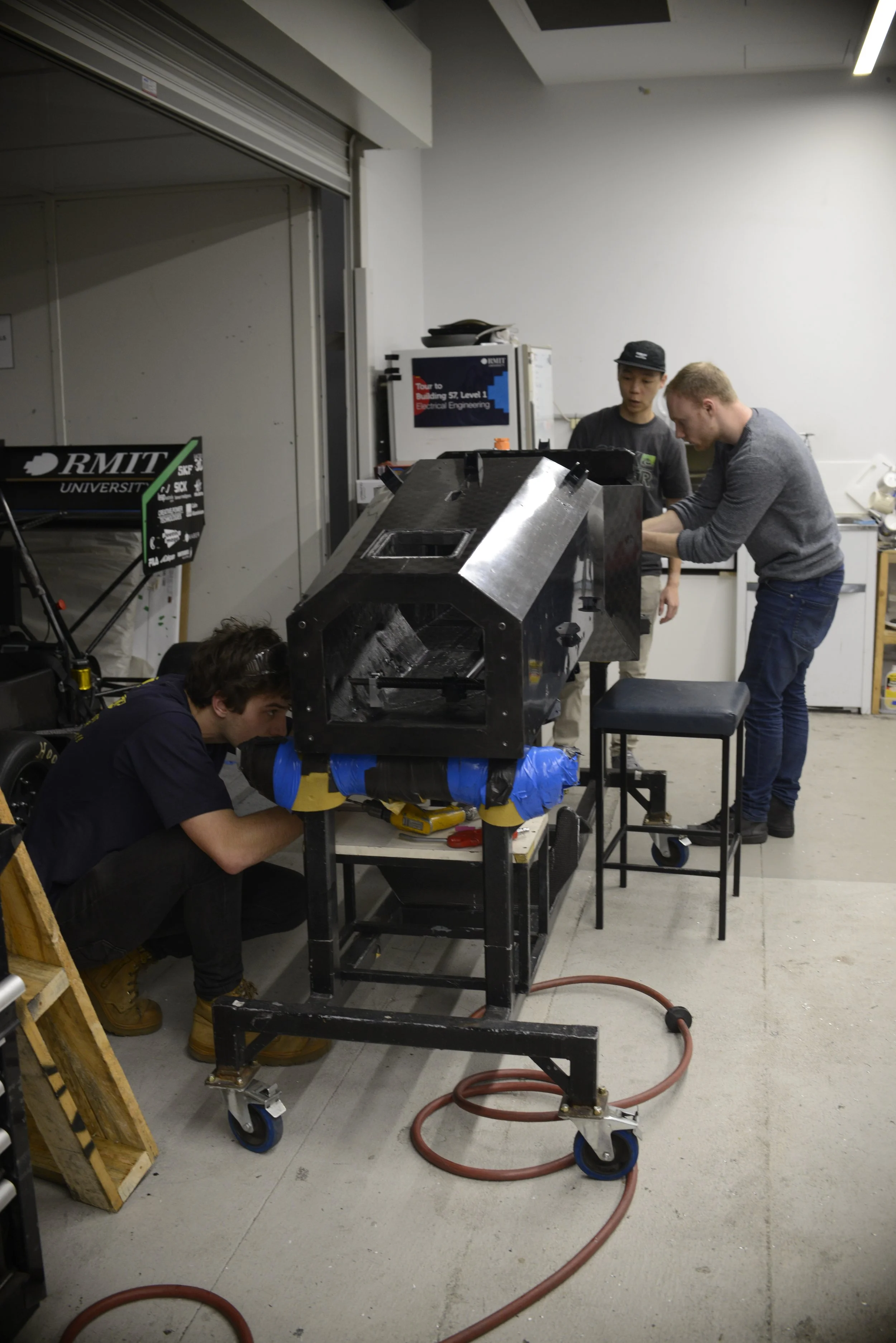 Three people working on a large black mechanical structure in a workshop, with university signage in the background.
