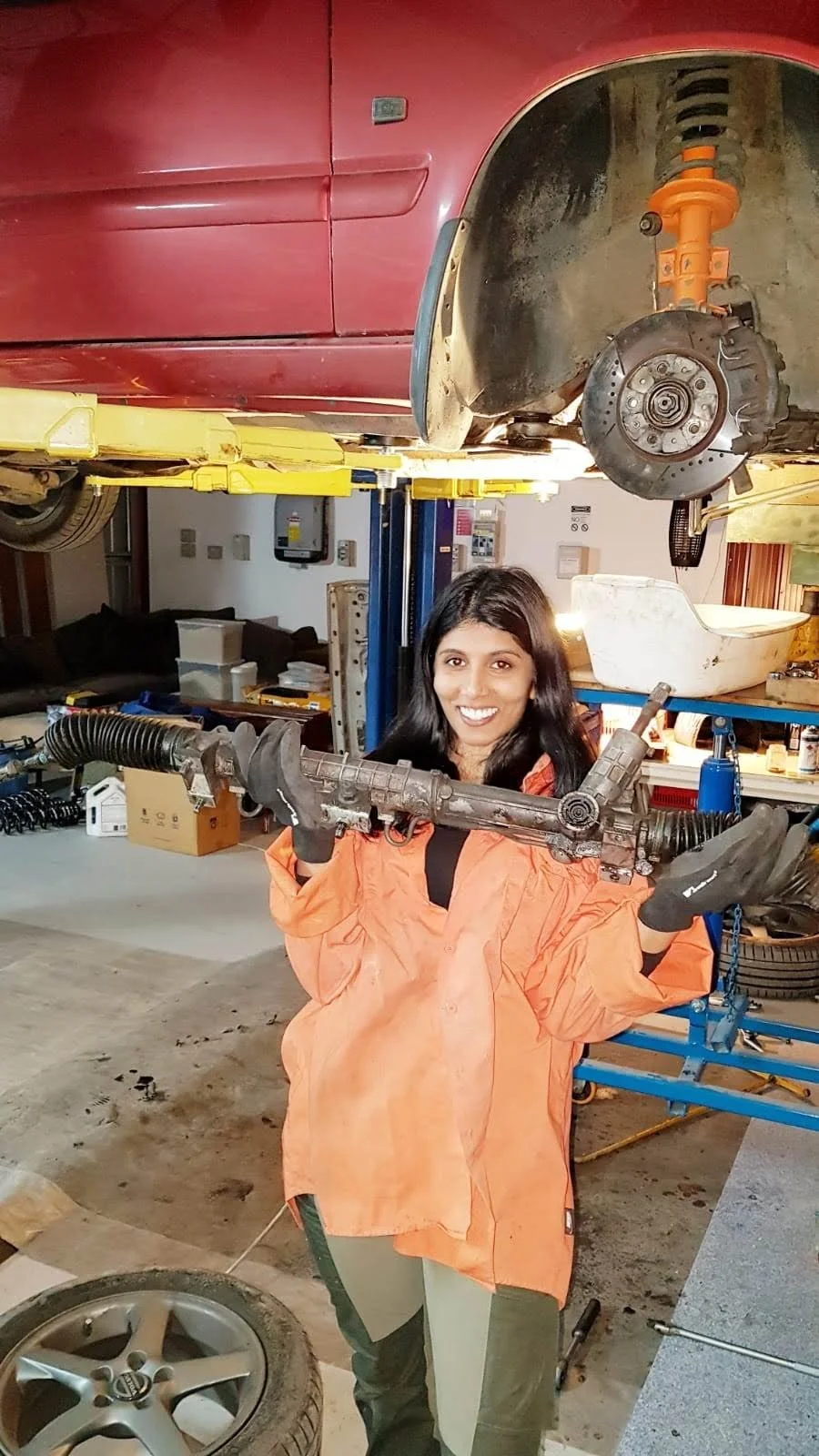 Woman in orange shirt holding a car part in a garage with a car on a lift; tire and tools visible.