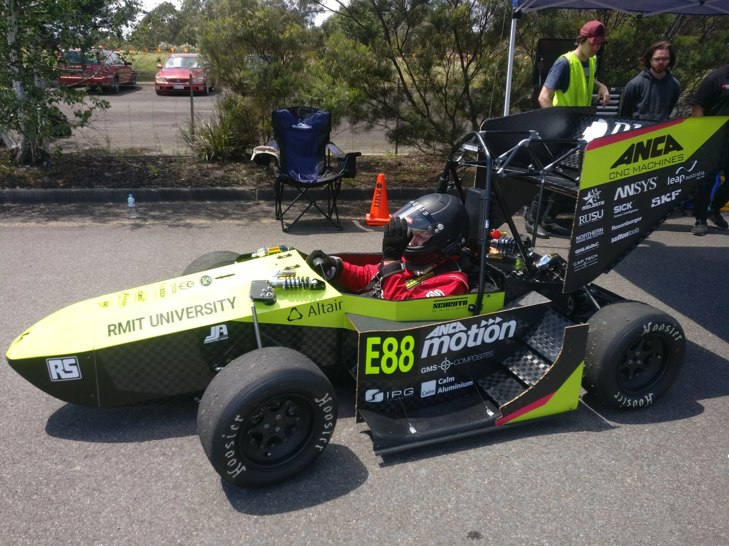 A driver in a racing suit and helmet sits in a green and black formula-style race car marked with "RMIT University" and various sponsor logos, parked on a road with people and cars in the background.