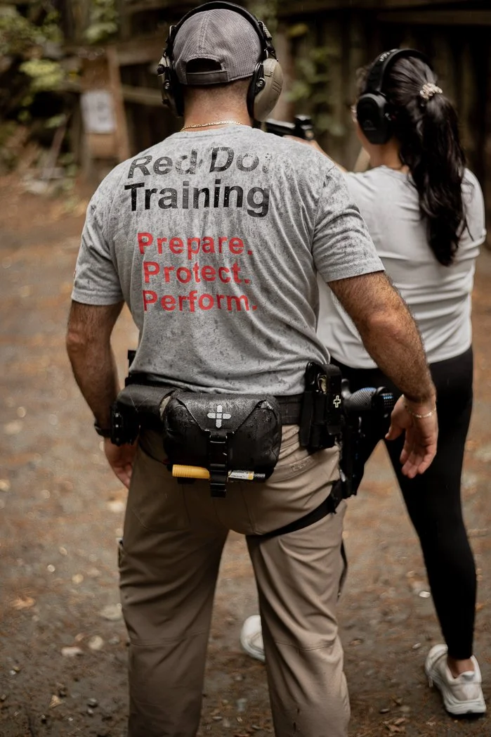 A man in a gray t-shirt and beige cargo pants with a firearm holstered at his waist, standing outdoors. The back of his shirt reads "Red Dot Training" and "Prepare. Protect. Perform." He is wearing a gray cap and headphones, with a woman next to him also wearing headphones, both participating in a firearm training session.