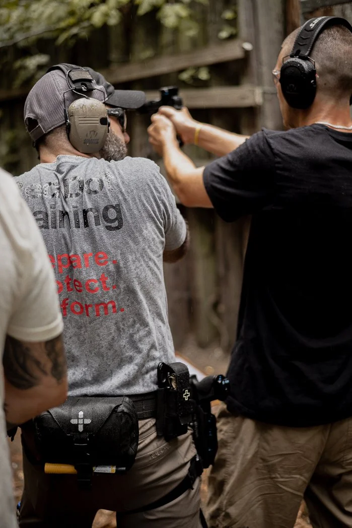 Two men at an outdoor shooting range, with one man aiming a handgun at a target while the other assists. Both are wearing ear protection, and one is in a gray t-shirt and the other in a black t-shirt.