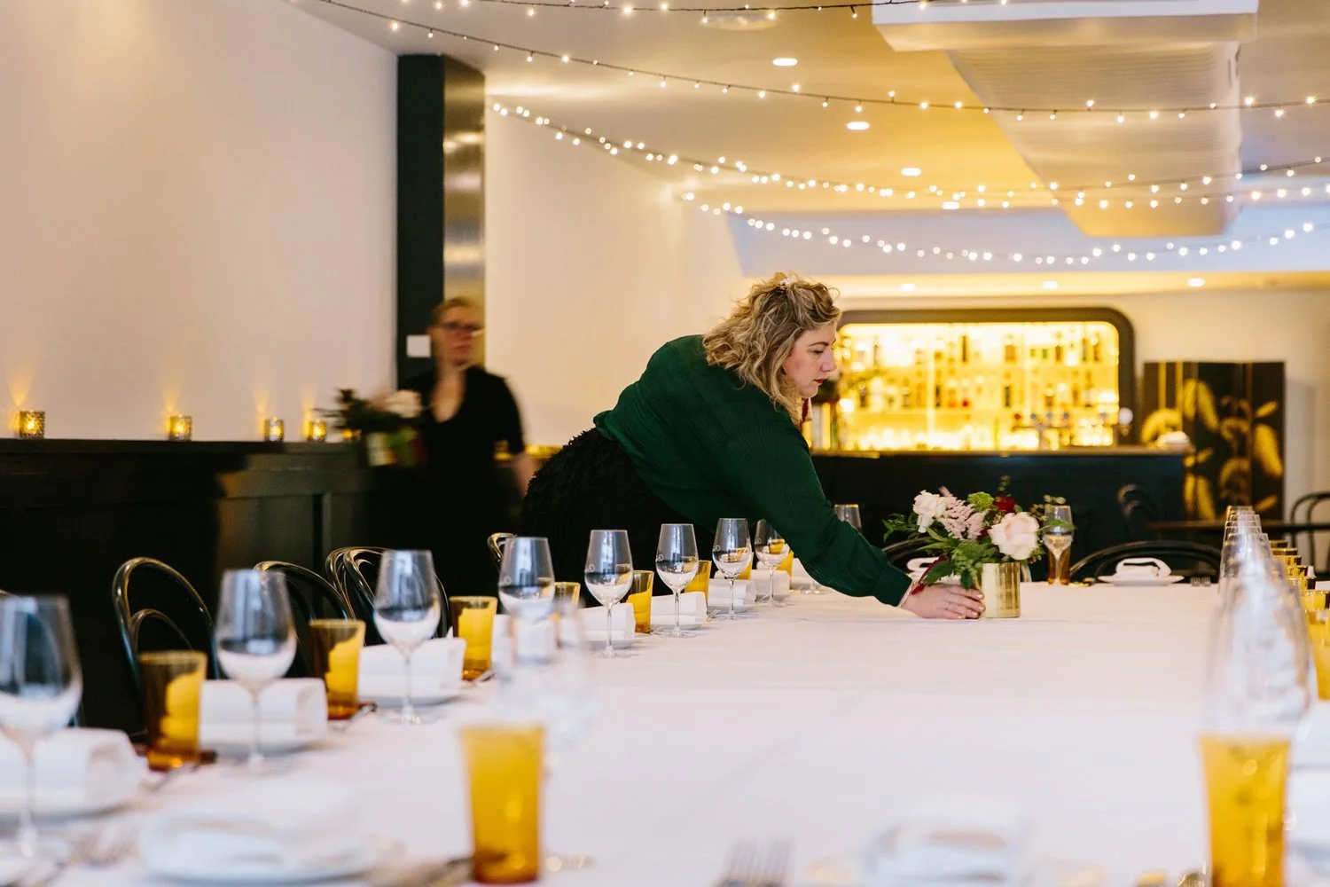A woman is setting a dinner table in a decorated restaurant or event space with a long white tablecloth, glassware, yellow cups, and a floral centerpiece, with string lights hanging overhead.
