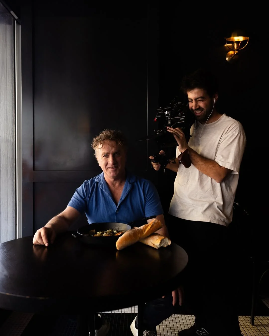 Adrian Richardson in a blue shirt sitting at a dark round table with a seafood dish and bread, while a Andrew Dorn films him inside a dimly lit restaurant or cafe.