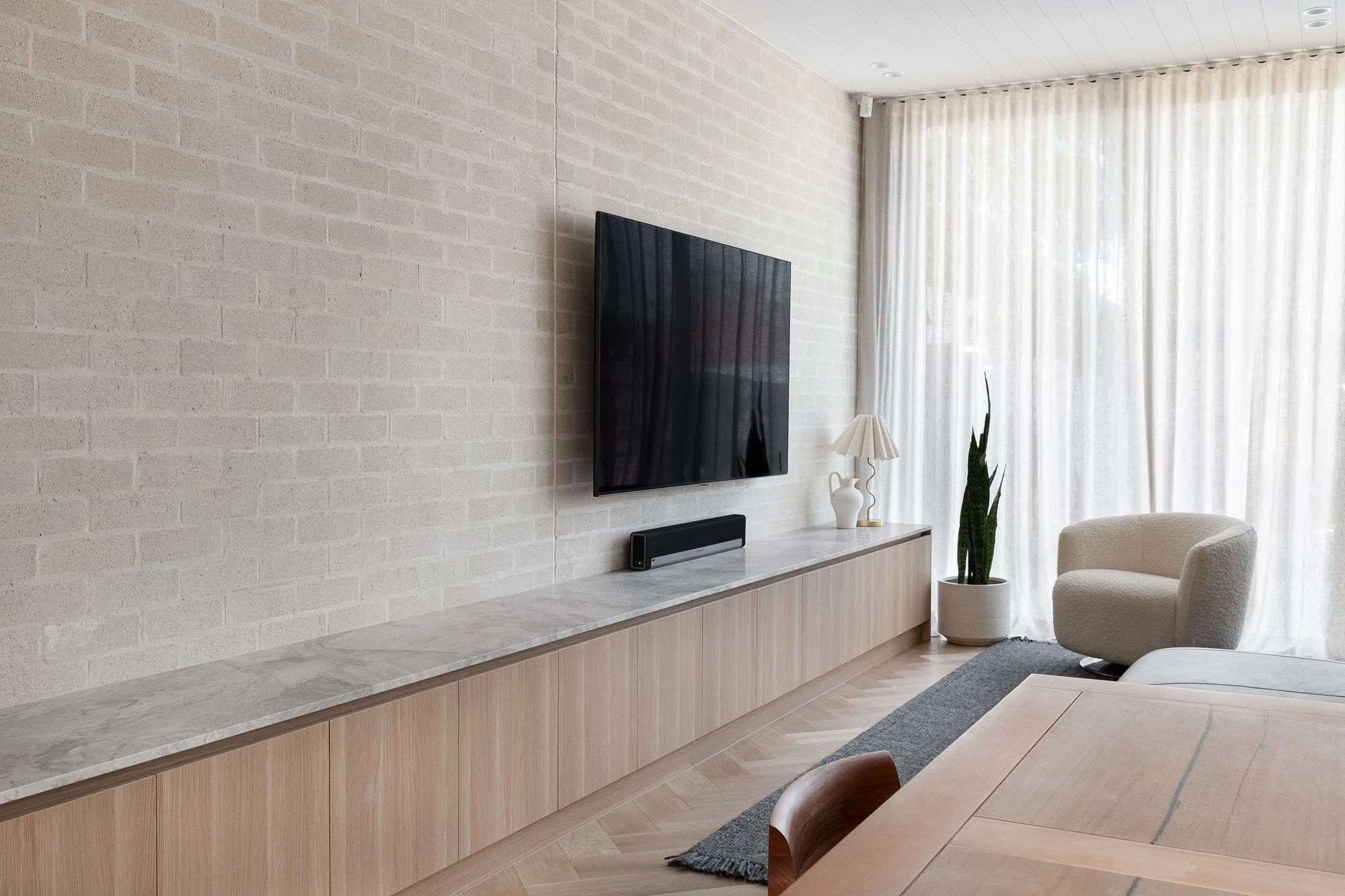 Modern living room with a wall-mounted flat-screen TV, white brick wall, white curtains, a beige armchair, a potted plant, a marble-top cabinet, and a wooden dining table.