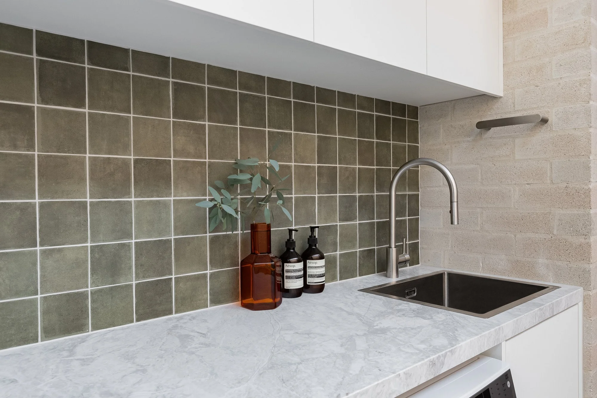 Kitchen countertop with a black sink, modern faucet, two brown bottles with black pump dispensers, and a brown vase with green plant, against a green tile backsplash.
