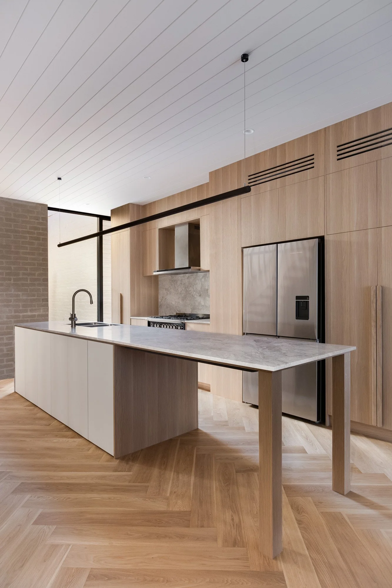 Modern kitchen with natural wood cabinets, marble countertop island, stainless steel refrigerator, and wooden flooring.
