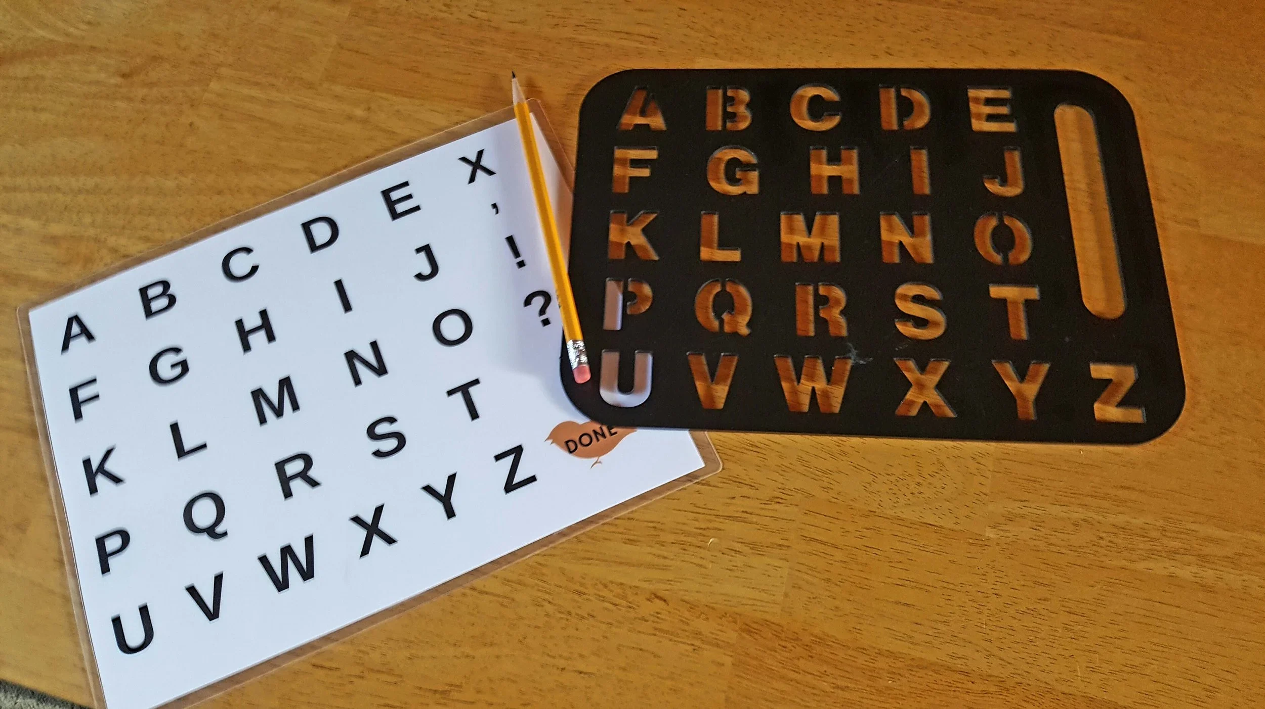 A black alphabet stencil with orange-brown letters, a white sheet with black uppercase alphabet letters, a yellow pencil, and a small paper cutout with the word 'DONE' on a wooden surface.
