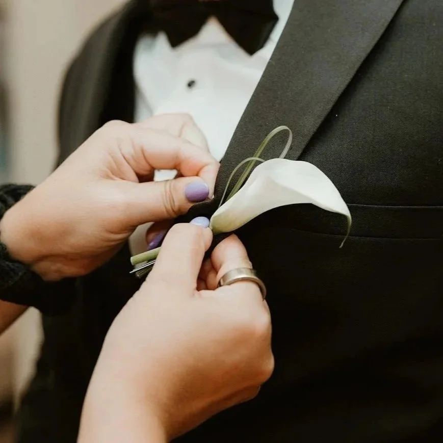 Someone is pinning a white calla lily flower to a man's black tuxedo jacket for a formal event.