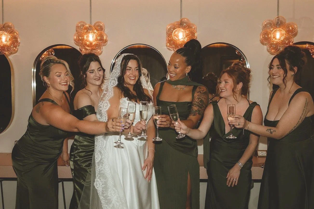 Group of women, some wearing bridesmaid dresses, celebrating and holding champagne glasses at a wedding reception.