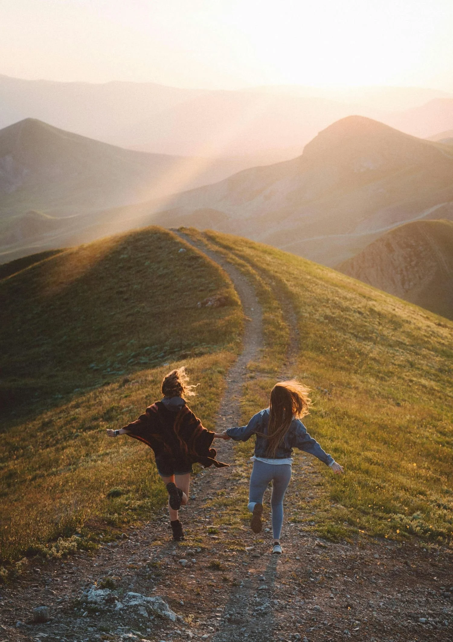 Two children holding hands and running down a dirt trail on a grassy hill during sunset, with rolling hills and mountains in the background.