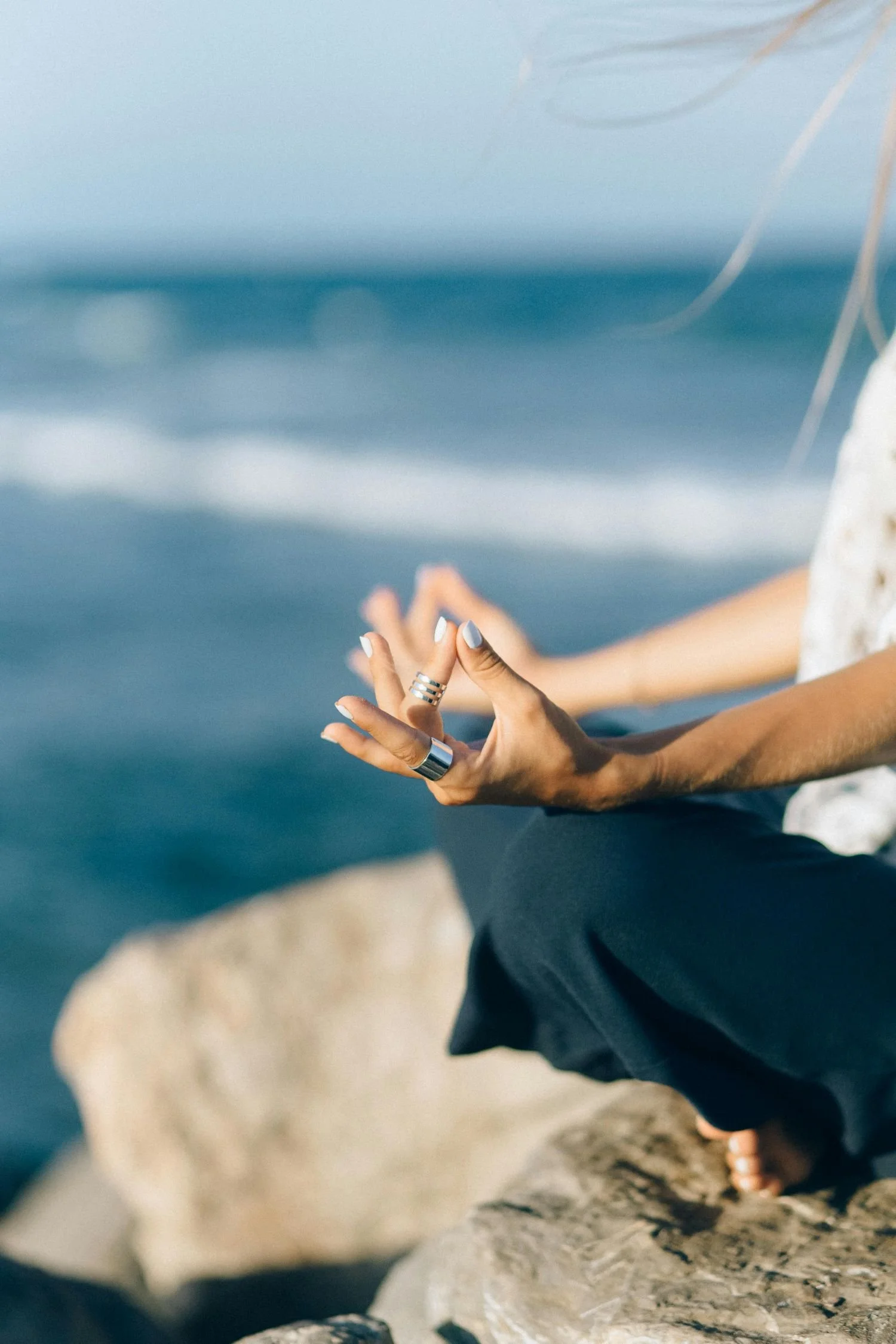Person practicing yoga or meditation on rocks by the ocean, focusing on their hands and legs.