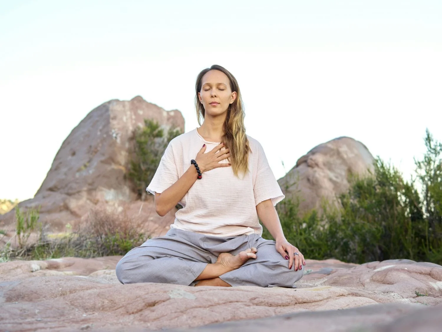 A woman practicing meditation outdoors, sitting cross-legged on rocks with her hand over her heart, eyes closed, and a calm expression.