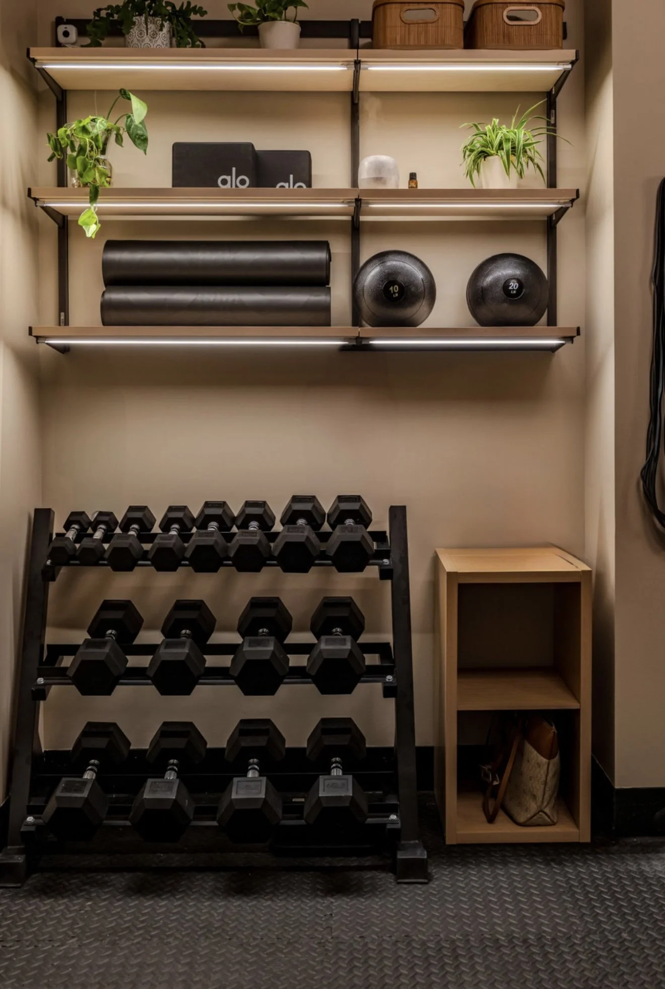 A home gym corner with shelves holding plants, workout equipment, and storage baskets, a rack with dumbbells, and a small wooden side table with a bag beneath it.